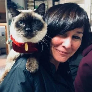 A woman with dark hair smiling next to a Siamese cat wearing a red collar with a gold tag.