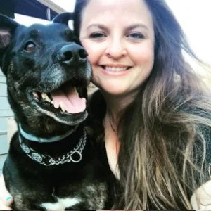 A woman with long hair smiling next to a black dog with a white chest, both looking happy.
