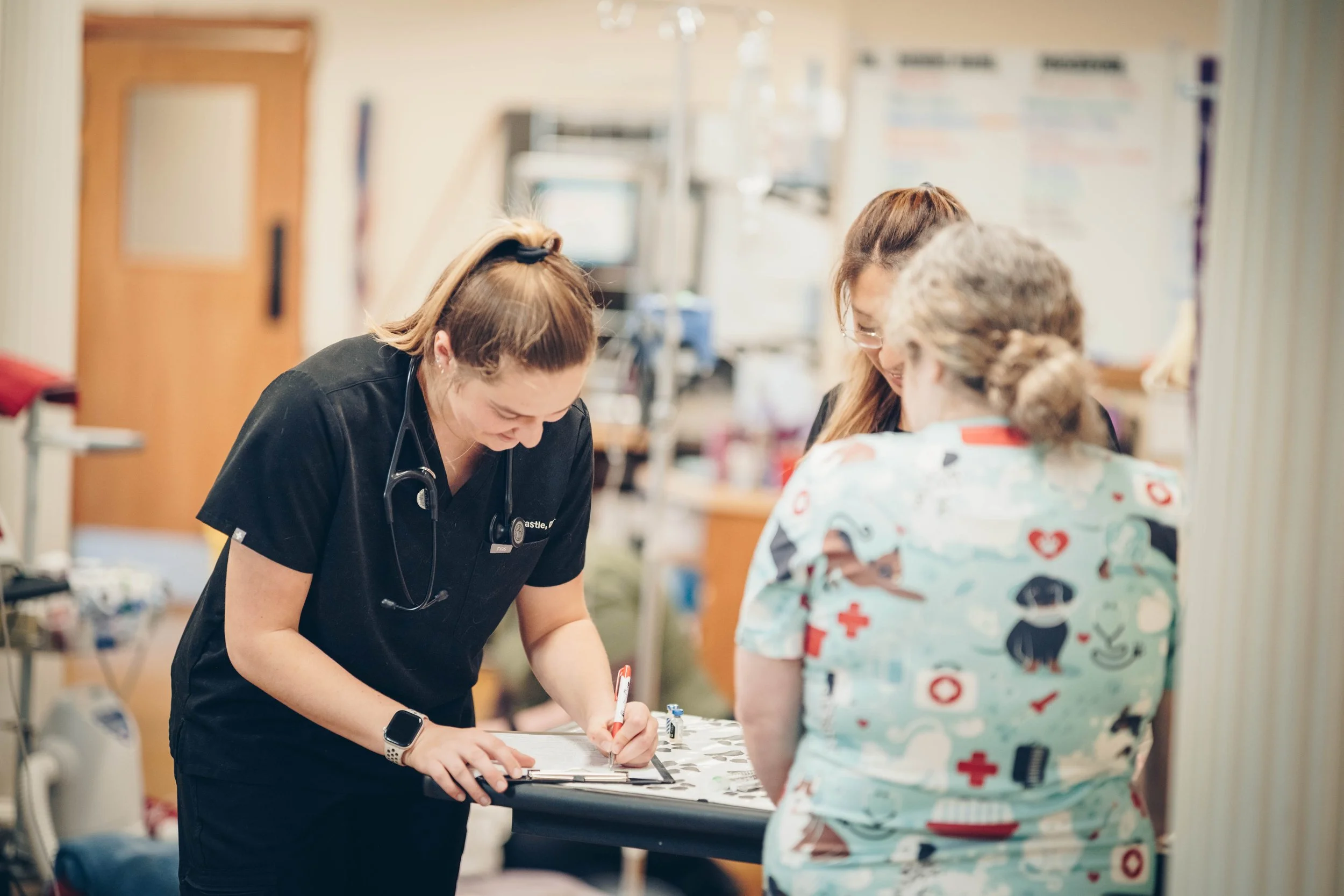 Three healthcare workers, including a nurse with a stethoscope, are gathered around a table in a hospital room, discussing and writing notes.