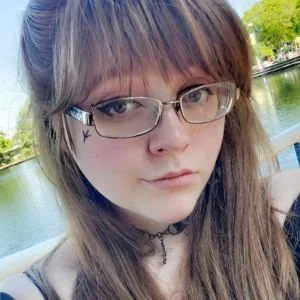 A young woman with long brown hair, glasses, and a choker necklace taking a selfie outdoors near a body of water, with trees in the background.