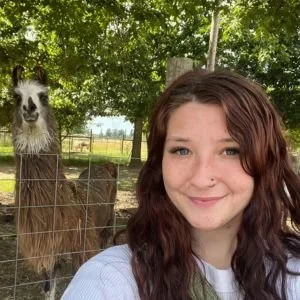 A girl with long, wavy, reddish-brown hair smiling in front of a fence with llamas behind it in a wooded outdoor setting.