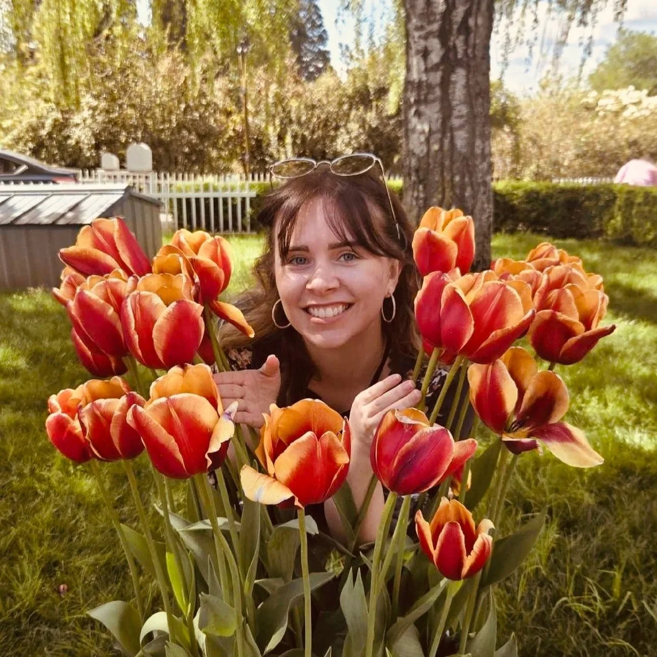 A woman with brown hair, smiling, wearing hoop earrings, squatting among orange-red tulips in a garden.