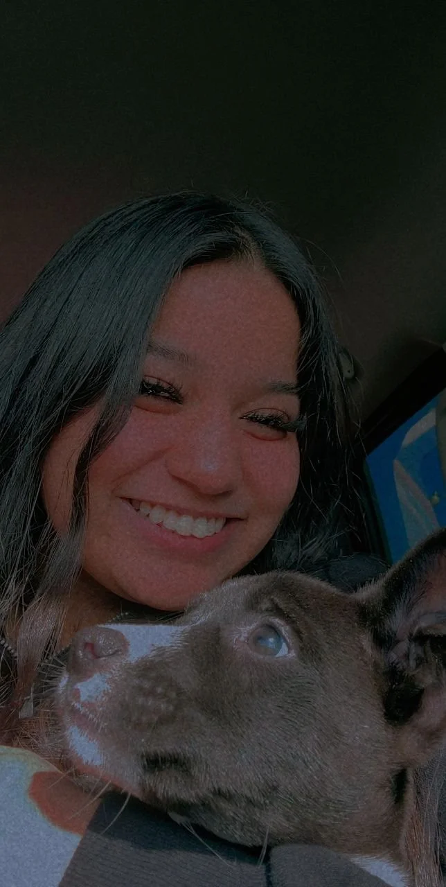 A woman with long dark hair smiling for a selfie with a brown puppy with white markings, inside a car.
