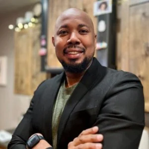 A smiling man with a beard and short hair standing with arms crossed in an indoor setting with wooden walls.