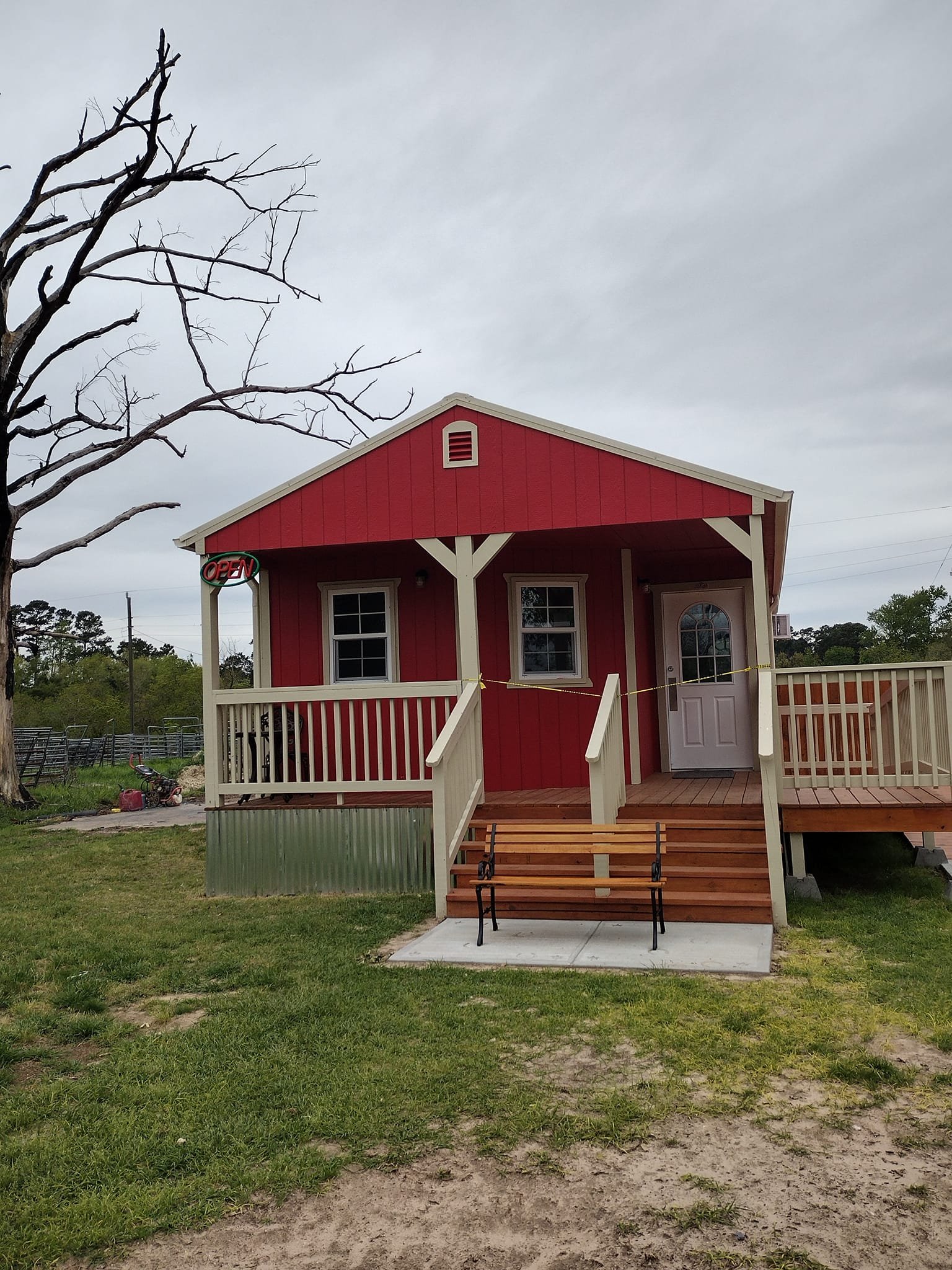 Red small house with white trim, front porch, stairs, and a bench in front, with a leafless tree on the left and cloudy sky overhead.