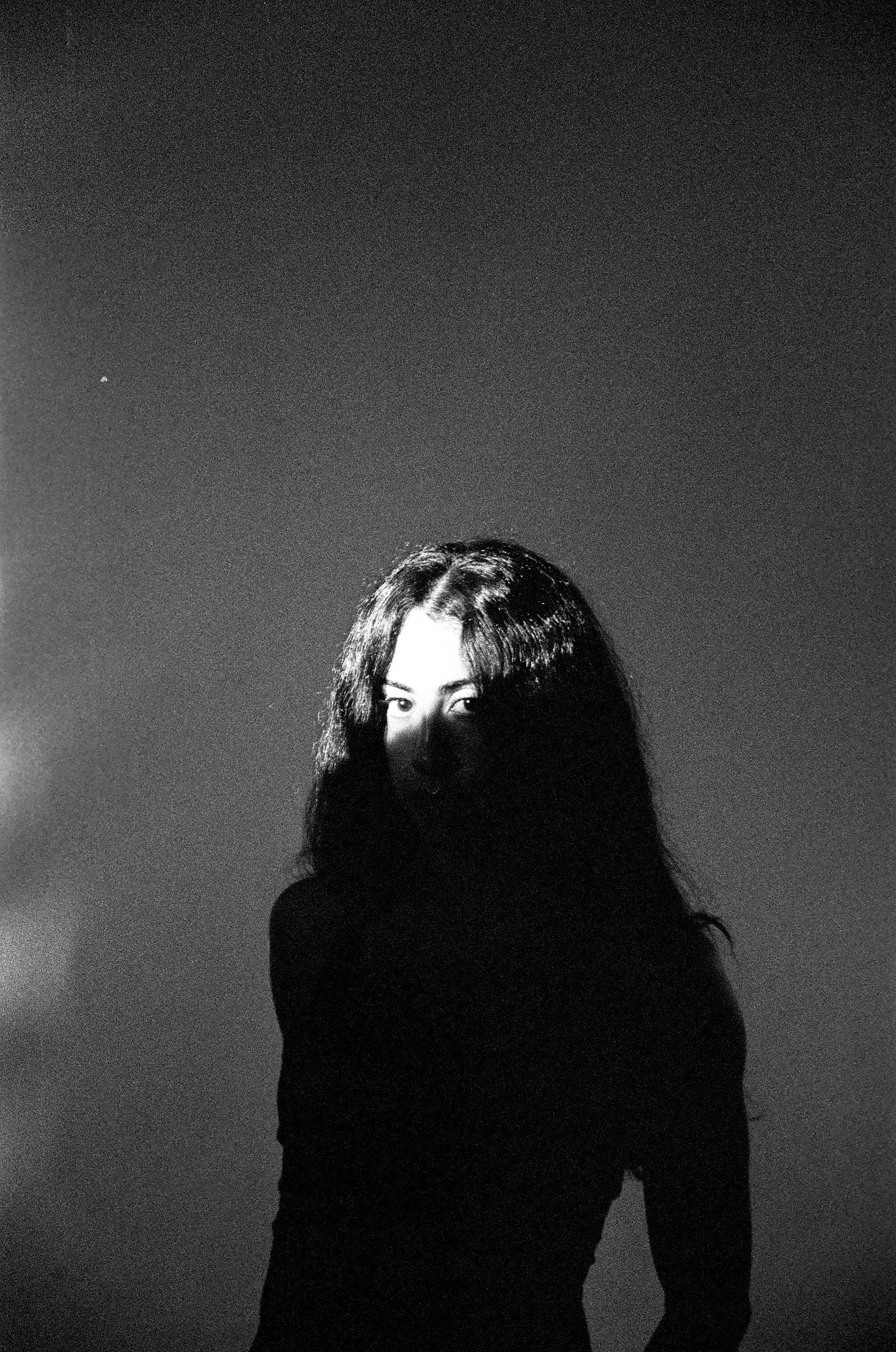 A woman stands in a room looking at the camera with all of her body in the dark except the top of her head.
