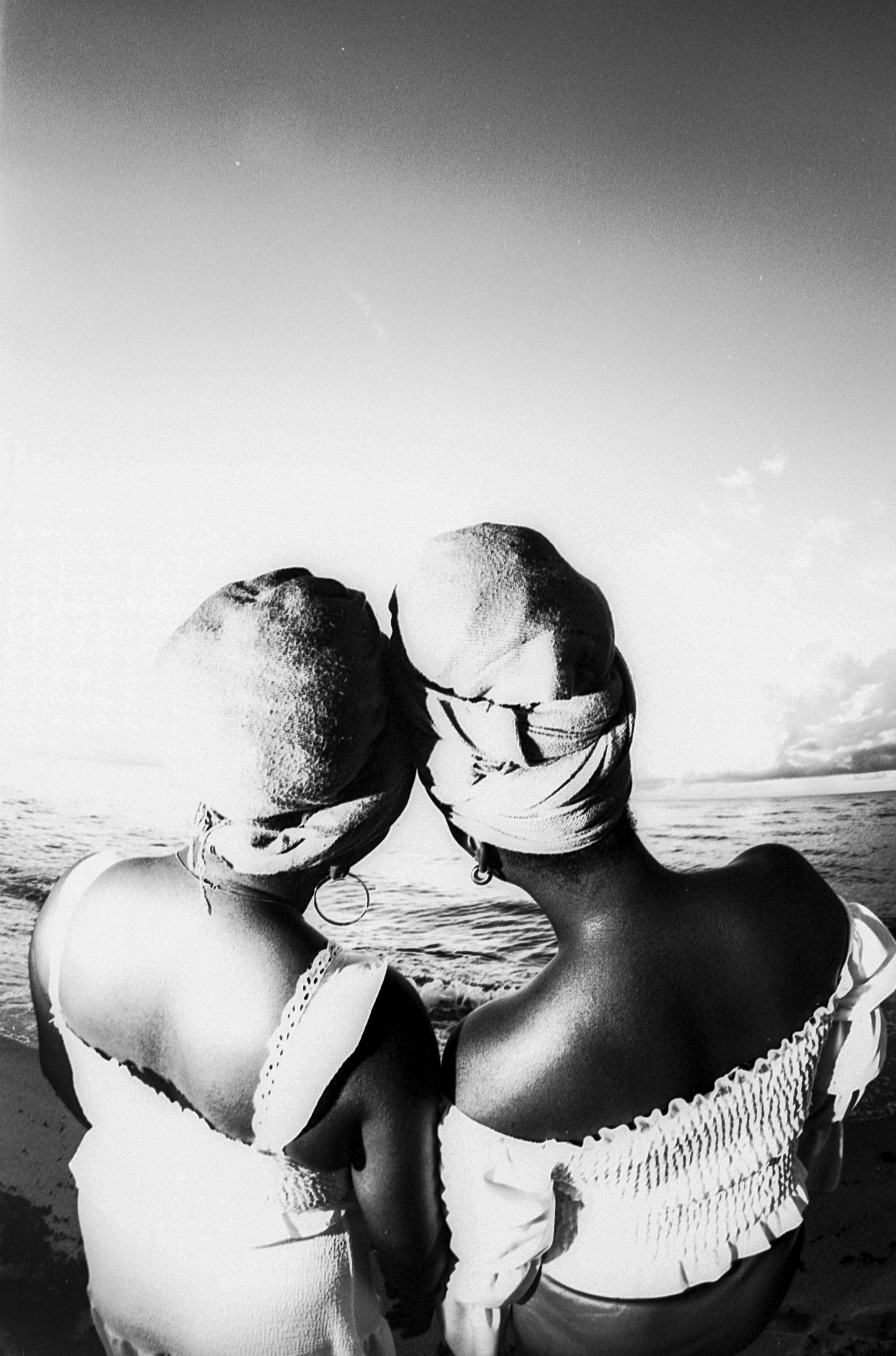 Two sisters with head wraps stand with their backs to the camera, looking out to the see with their heads tilted towards one another.