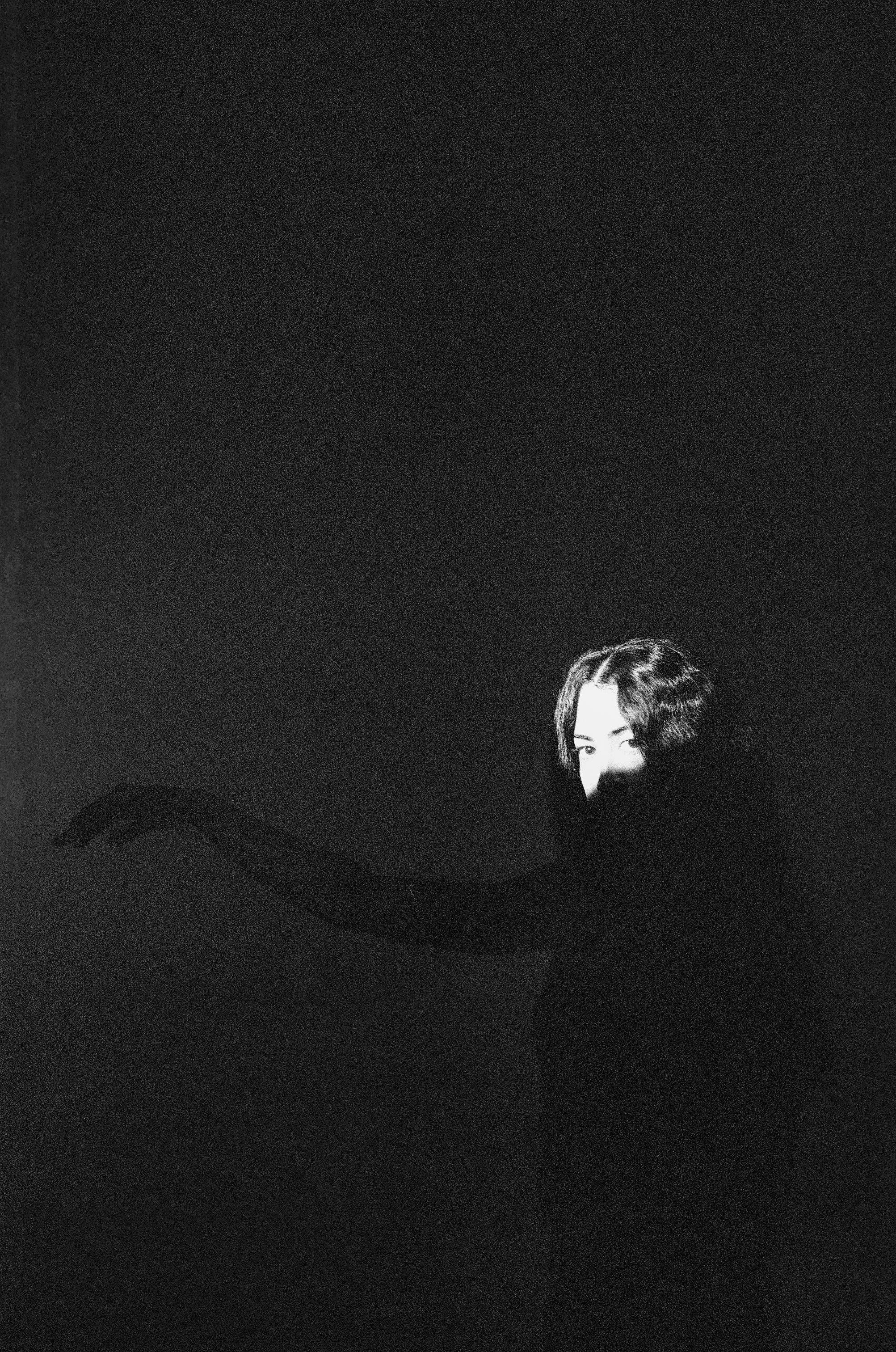 A woman stands in a room looking at the camera with all of her body in the dark except the top of her head.