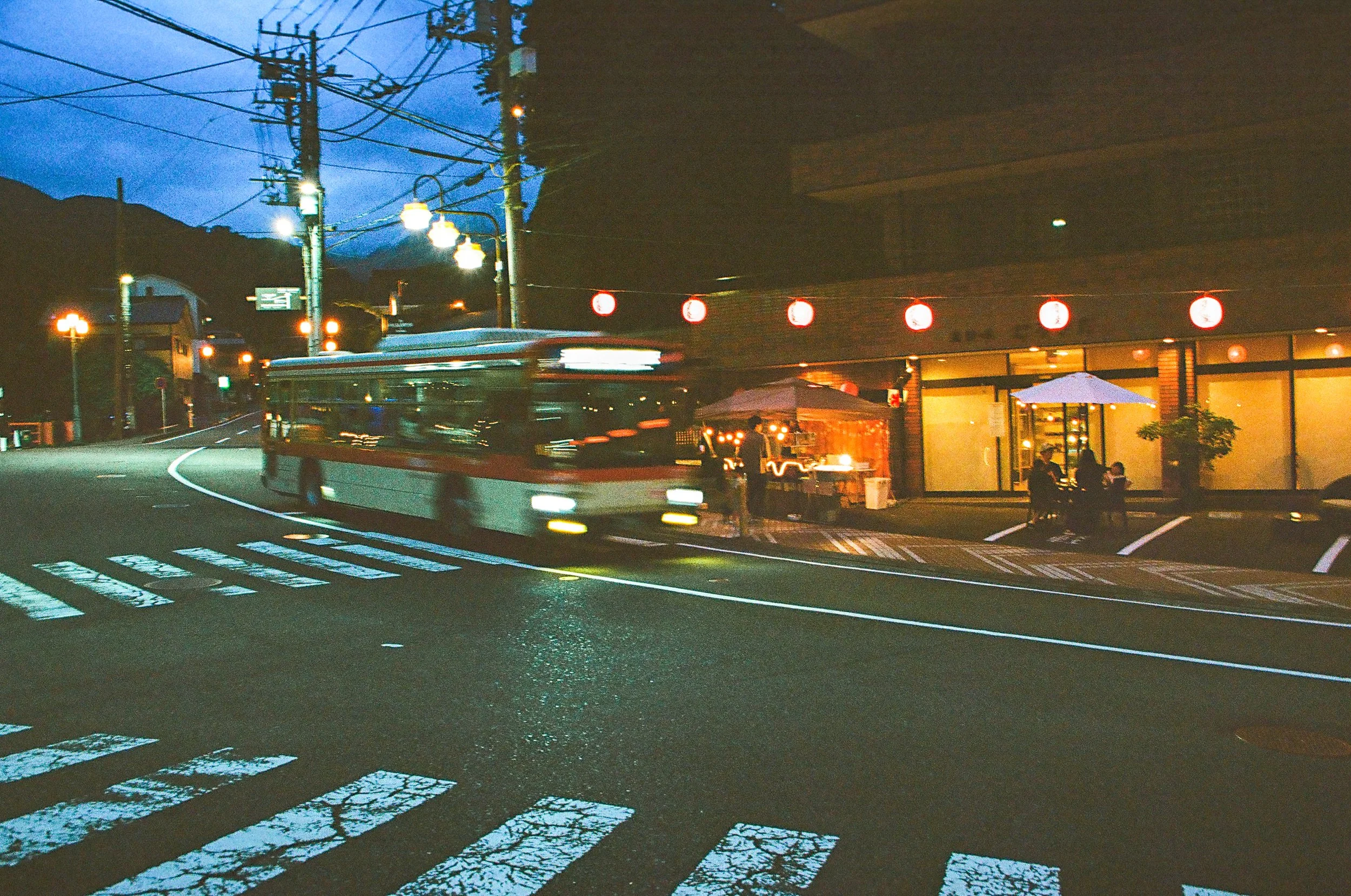 A city street scene at dusk with a moving bus, illuminated street lamps, and a building with red lanterns, against a backdrop of mountains.