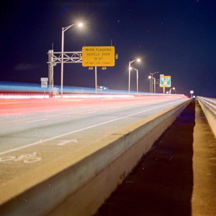 A highway at night with light trails from moving vehicles, streetlights illuminating the road, and a yellow traffic sign indicating conditions for flashing vehicle over 15 feet.