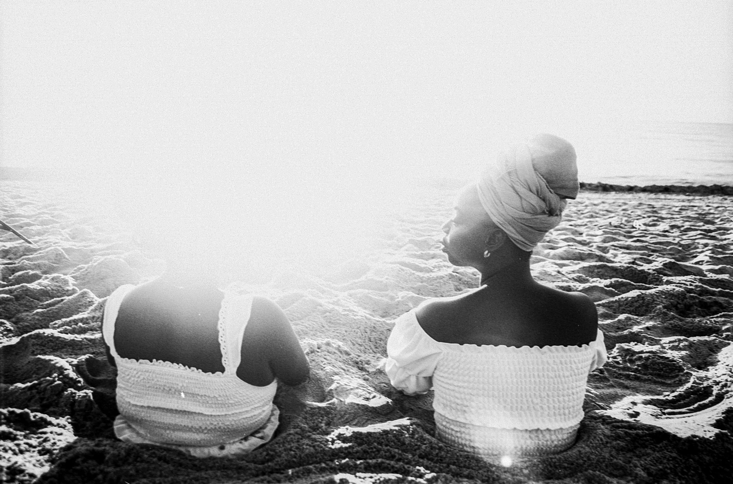 Two sisters sit in sand on either side of each other and are looking at each other. The glare of the sun covers the woman on the left side.