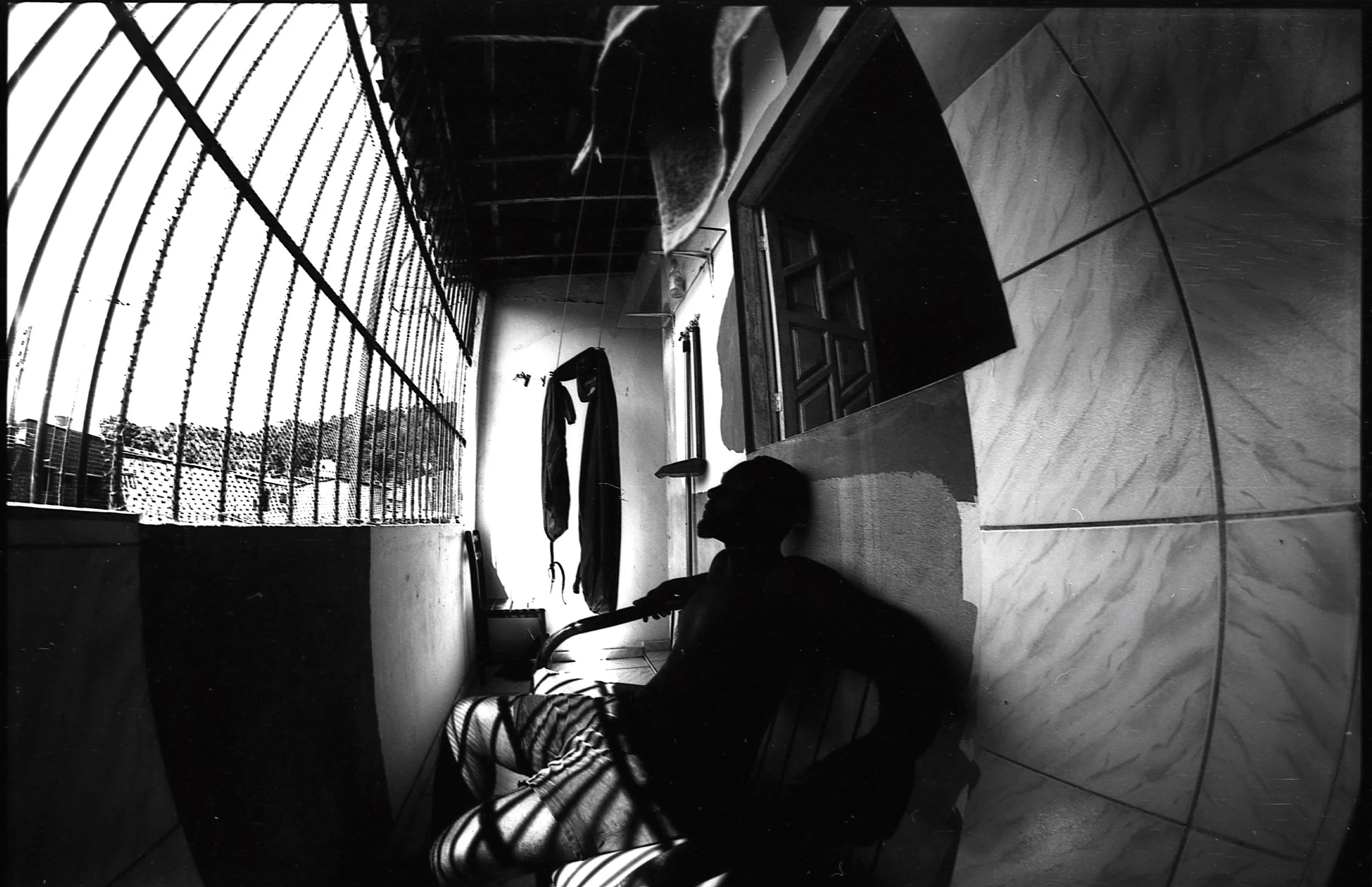 A person sitting on a bench in a room with marble tile flooring, facing a window with metal bars, with clothes hanging on a clothesline and a desk with a drawer, all in black and white.