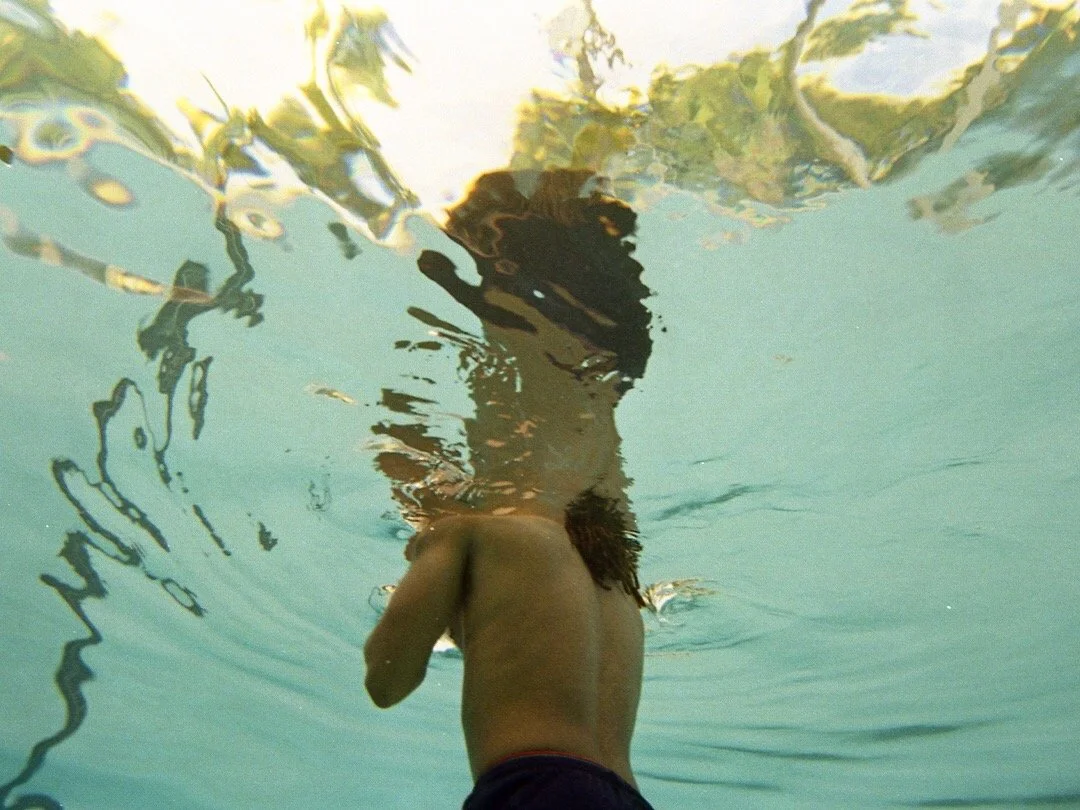 Underwater view of a man with long hair, shirtless, in a swimming pool, with rippling water and reflections on the surface.
