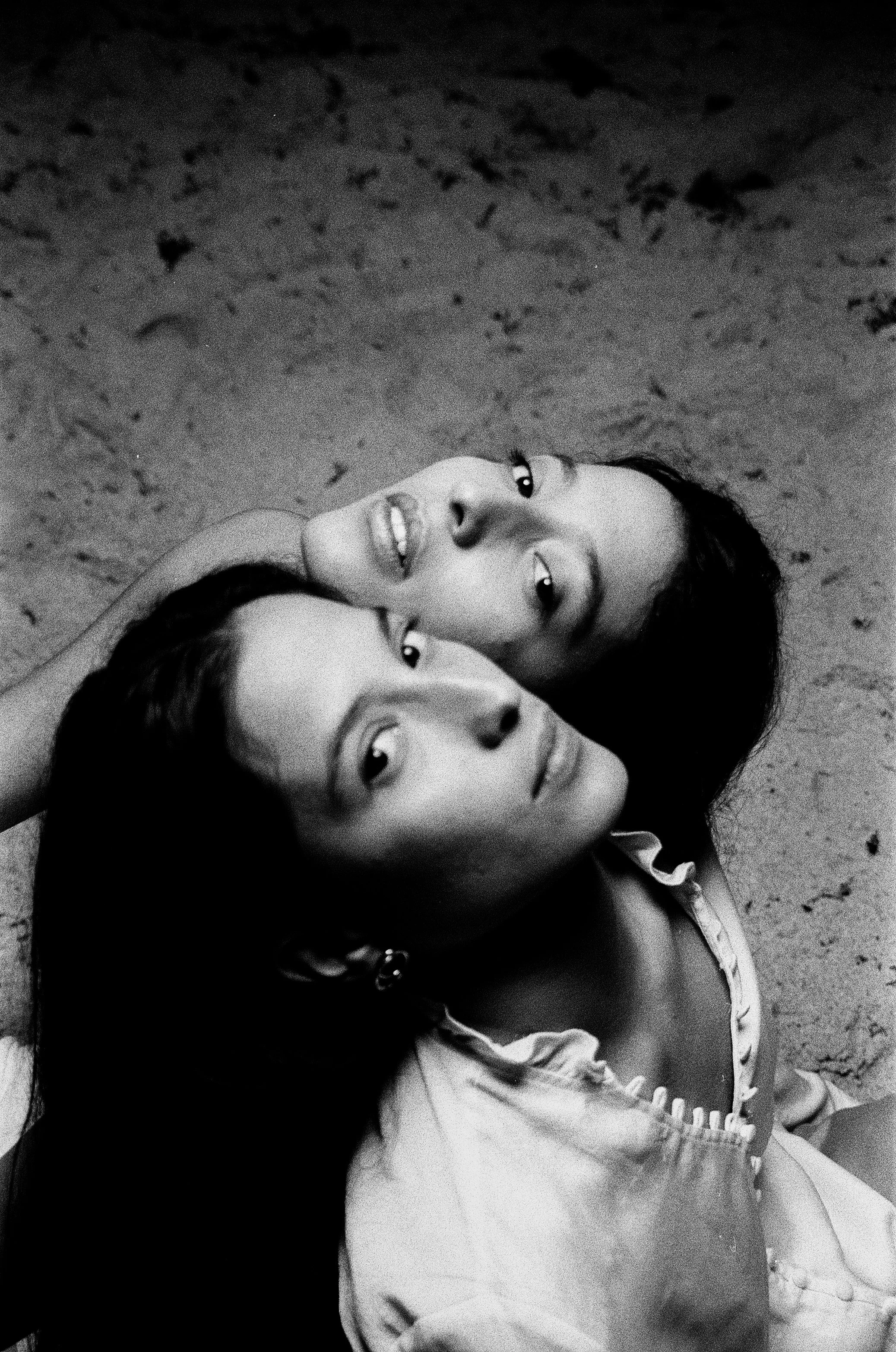 Two sisters with long hair are seated in sand, looking up towards the camera with their faces pressed together.