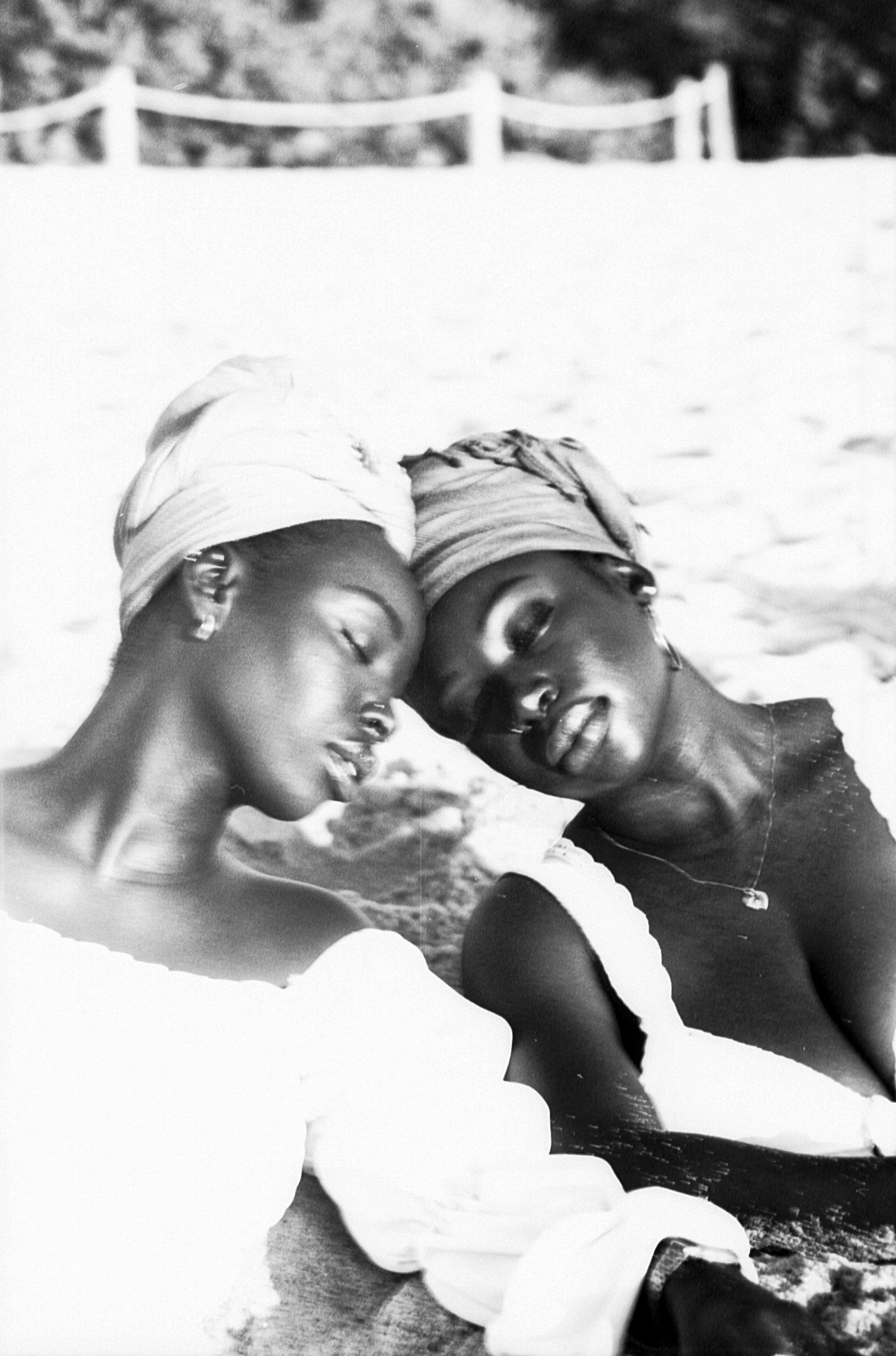 Two sisters with beautiful head wraps sit in sand with eyes closed with their heads touching.