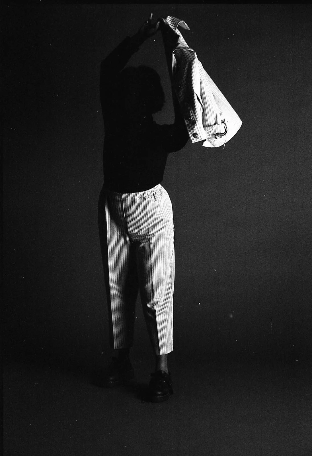 A full body image of a woman standing with her jacket held over the right side of her upper body. The jacket casts a shadow over her face because the light was coming from the right side.