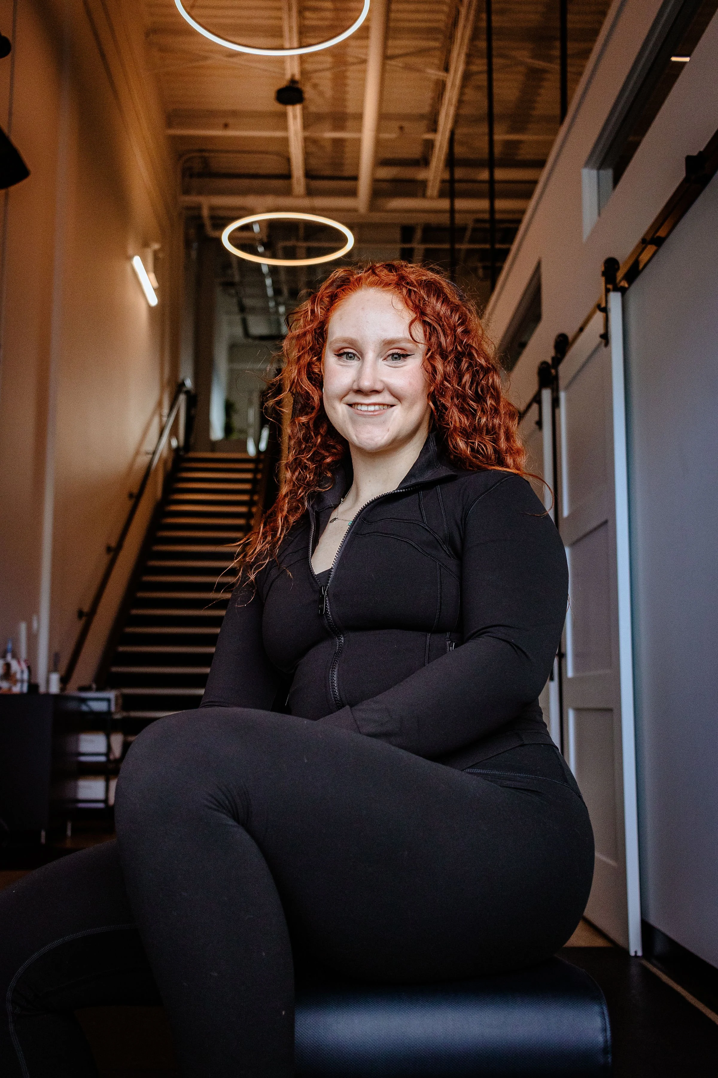 A young woman with curly red hair, smiling, sitting cross-legged in a modern, industrial-style interior with wooden ceiling and black metal stairs.