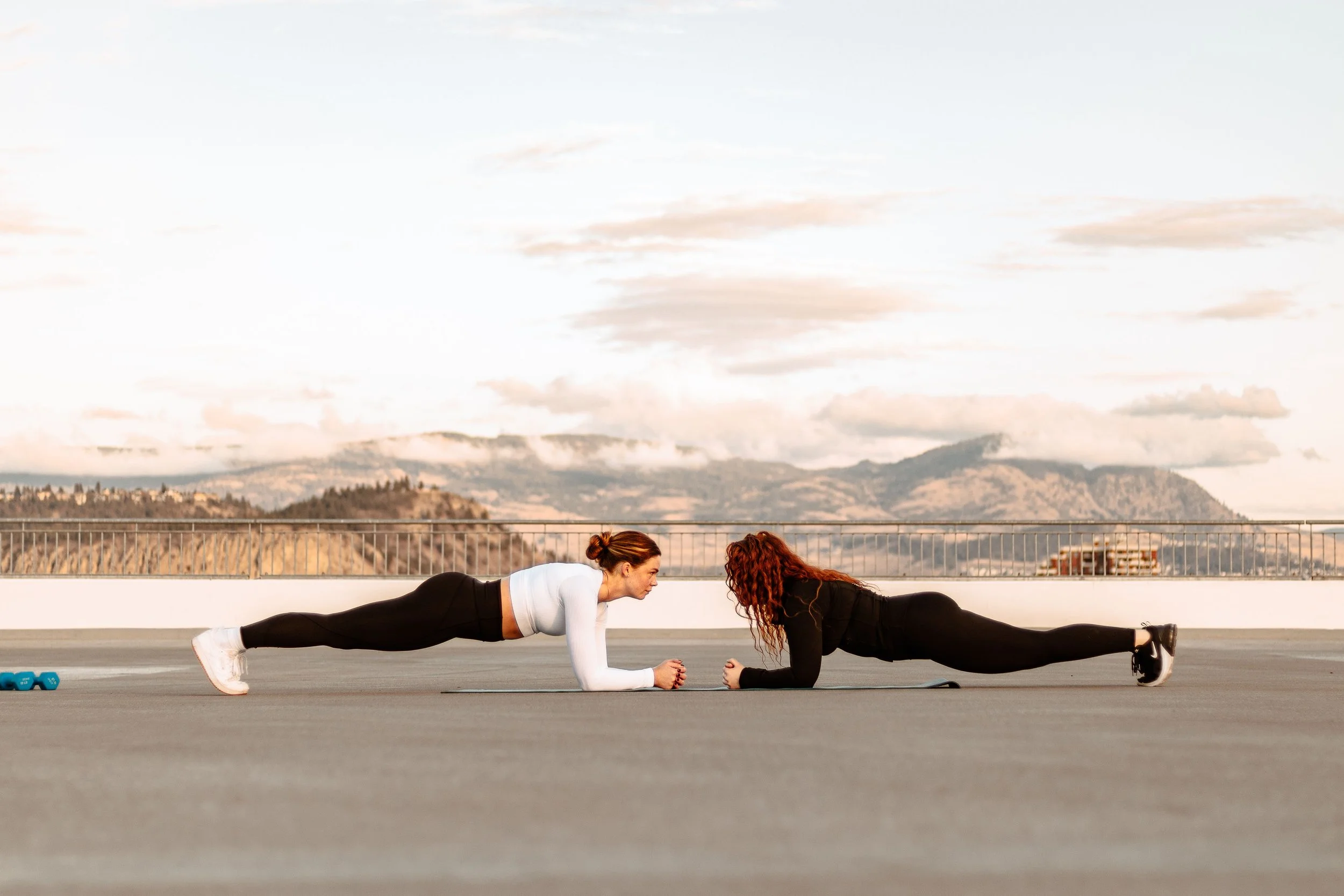 Two women exercising outdoors on a rooftop, performing planks facing each other with mountains in the background.