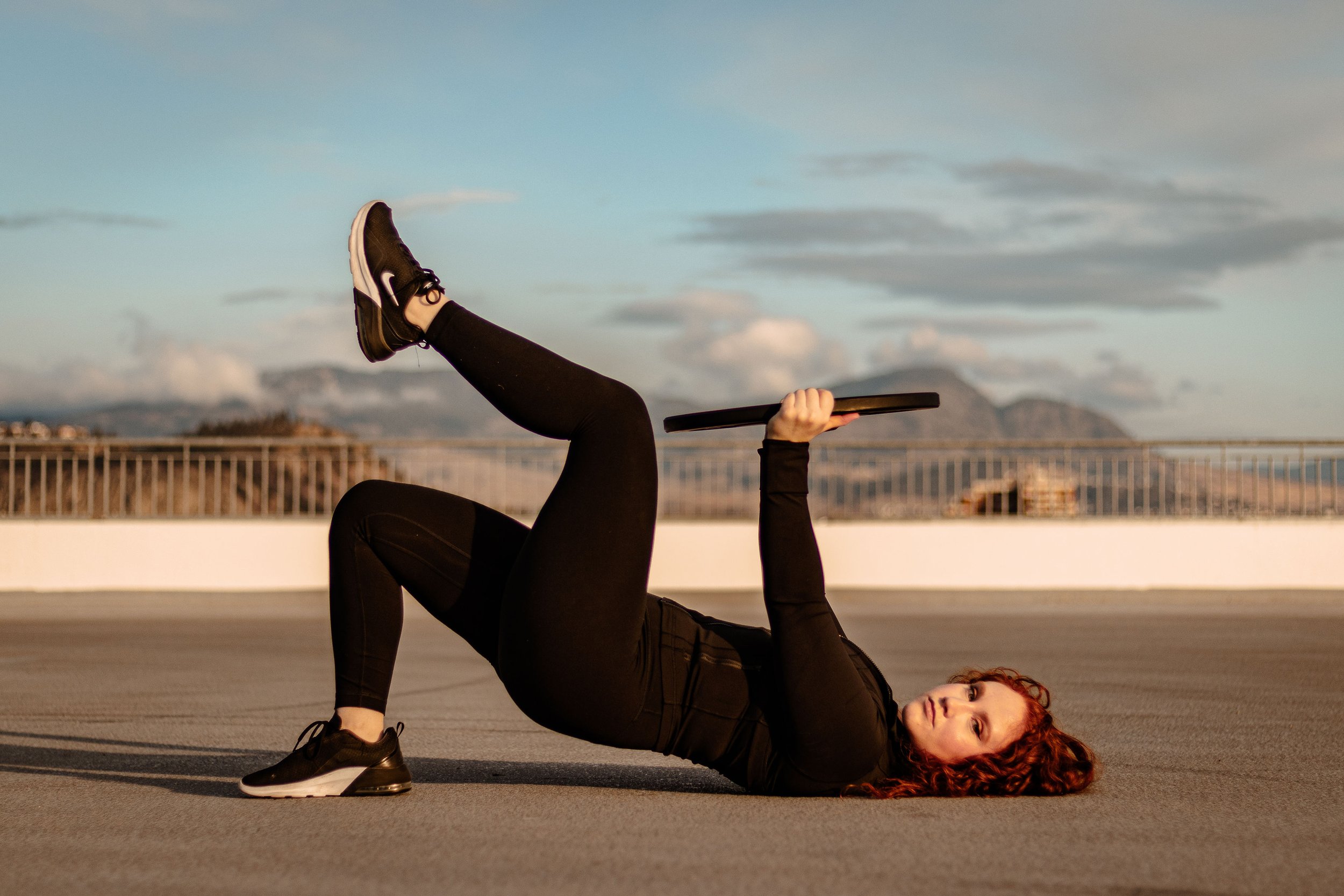 Woman lying on her back on a rooftop, holding a weight plate, performing a glute bridge exercise, with mountains and a partly cloudy sky in the background.