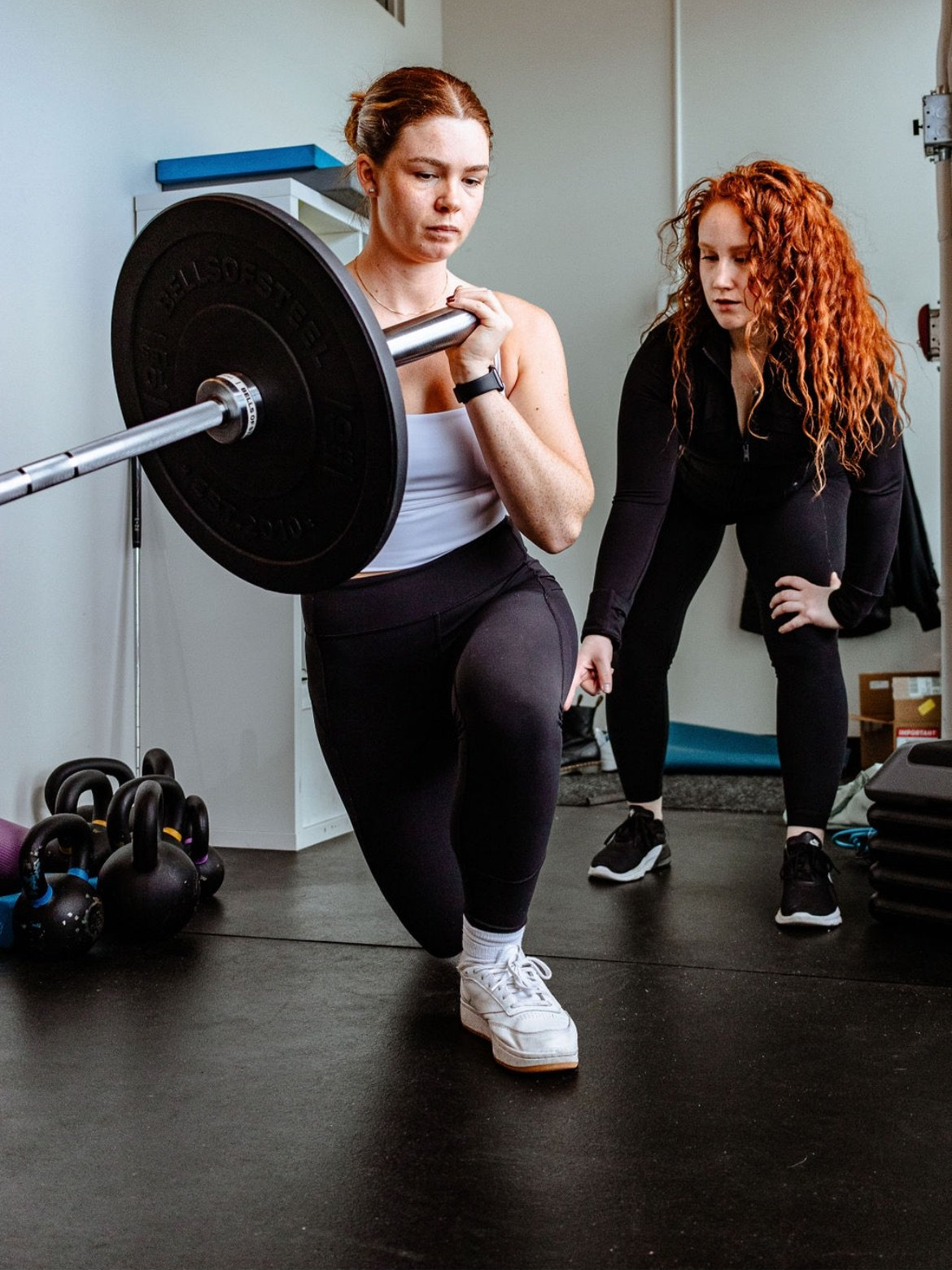 A woman is lifting a barbell in a gym while a trainer guides her. The woman has red hair and is wearing black leggings and a white tank top. The trainer, with curly red hair, is wearing black workout clothes and is observing closely.