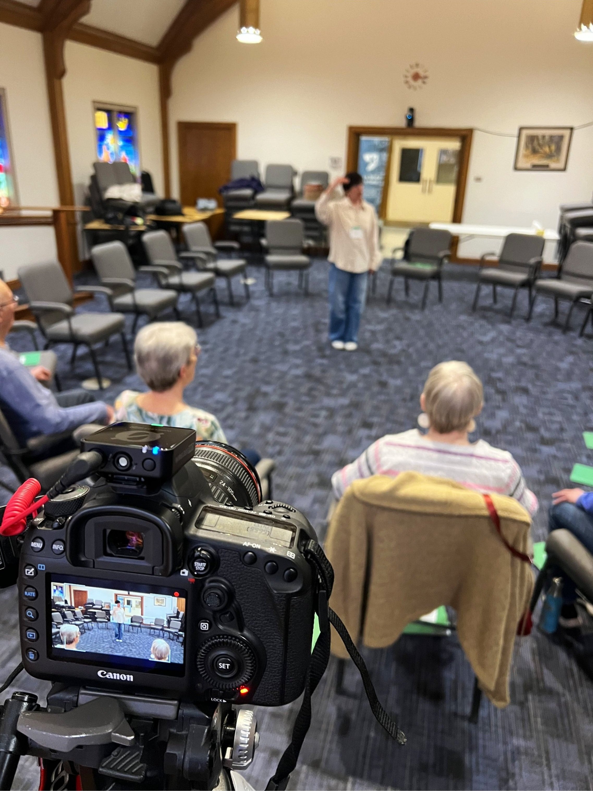 A woman standing in front of seated audience in a meeting room, with a camera capturing the scene.