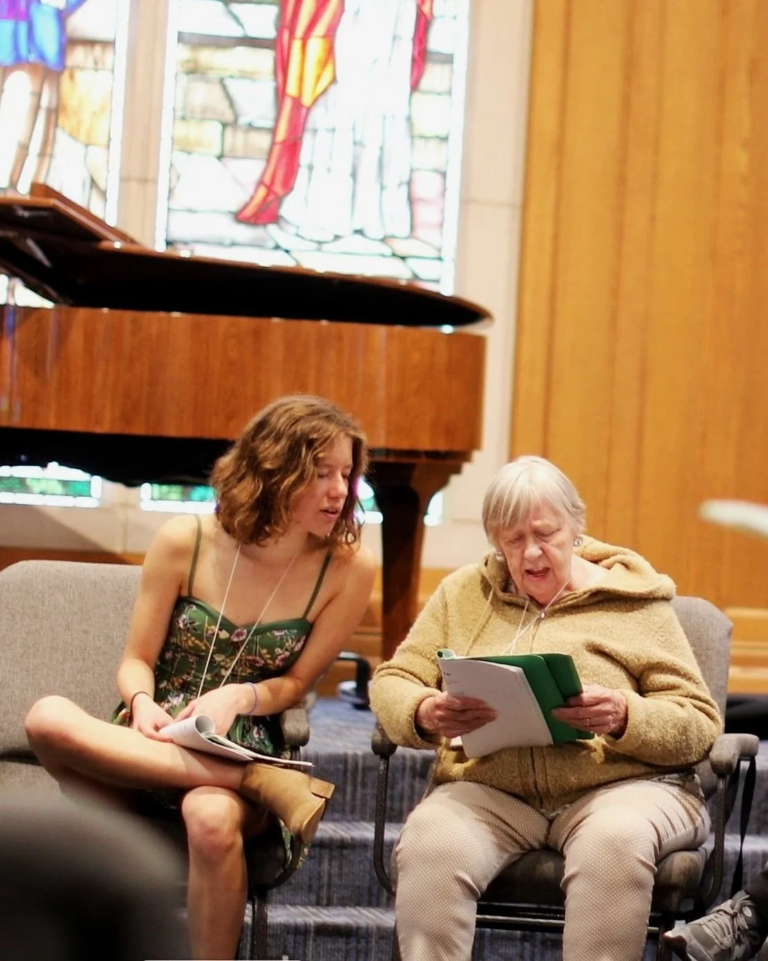 Two women sitting on chairs in front of a grand piano, reading sheet music and singing, with a stained glass window in the background.