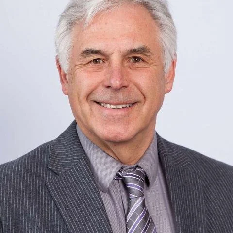 A smiling middle-aged man with gray hair, wearing a dark pinstripe suit, a light gray shirt, and a striped purple and gray tie, standing against a plain light background.