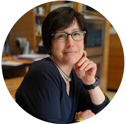 A woman with short dark hair, glasses, and a navy blue top sitting at a desk in an office, resting her chin on her hand and smiling.