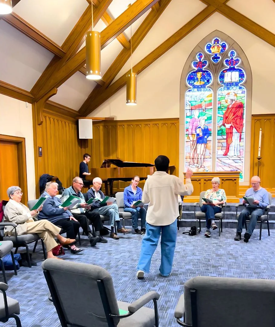 A group of people seated in a semi-circle inside a church, singing from songbooks, with a conductor standing in front of them. A pianist plays on a grand piano, and a stained-glass window depicting biblical scenes is in the background.