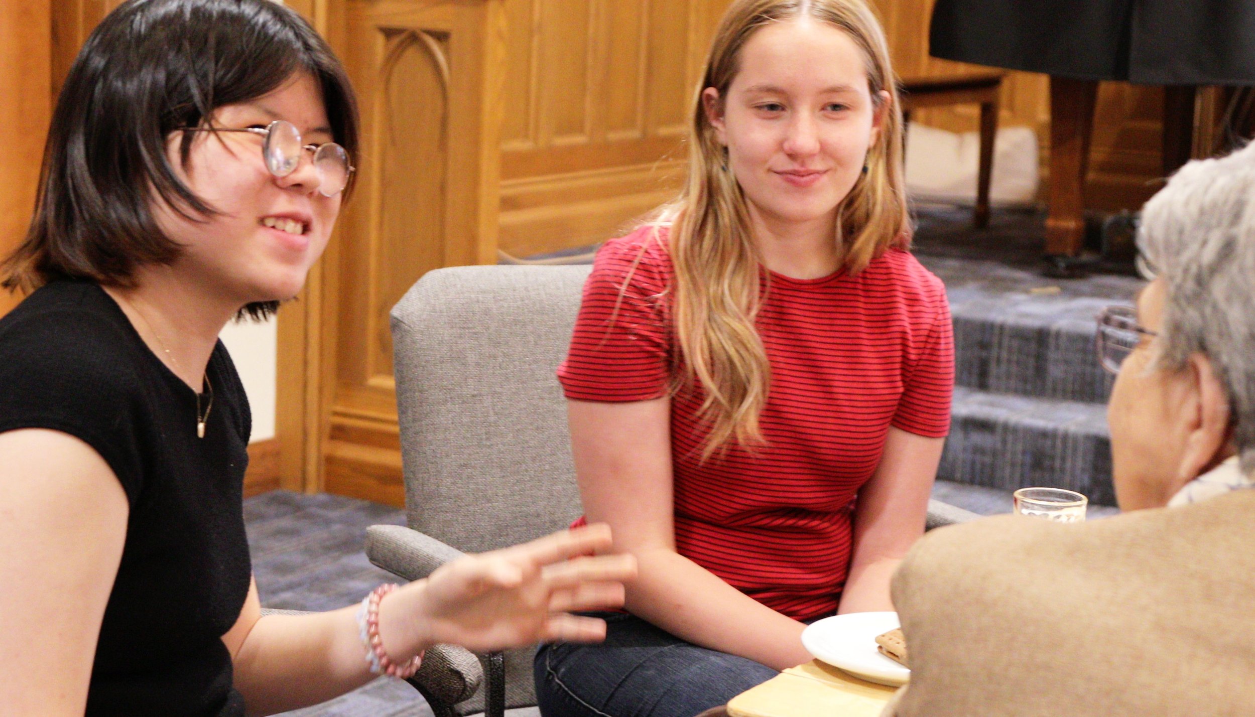 Three women are sitting at a table having a conversation. The woman on the left has short dark hair, glasses, and wears a black top. The woman in the middle has long blonde hair and wears a red striped shirt. The woman on the right has gray hair, glasses, and wears a beige jacket. There are plates and a glass on the table, and a wooden interior background.