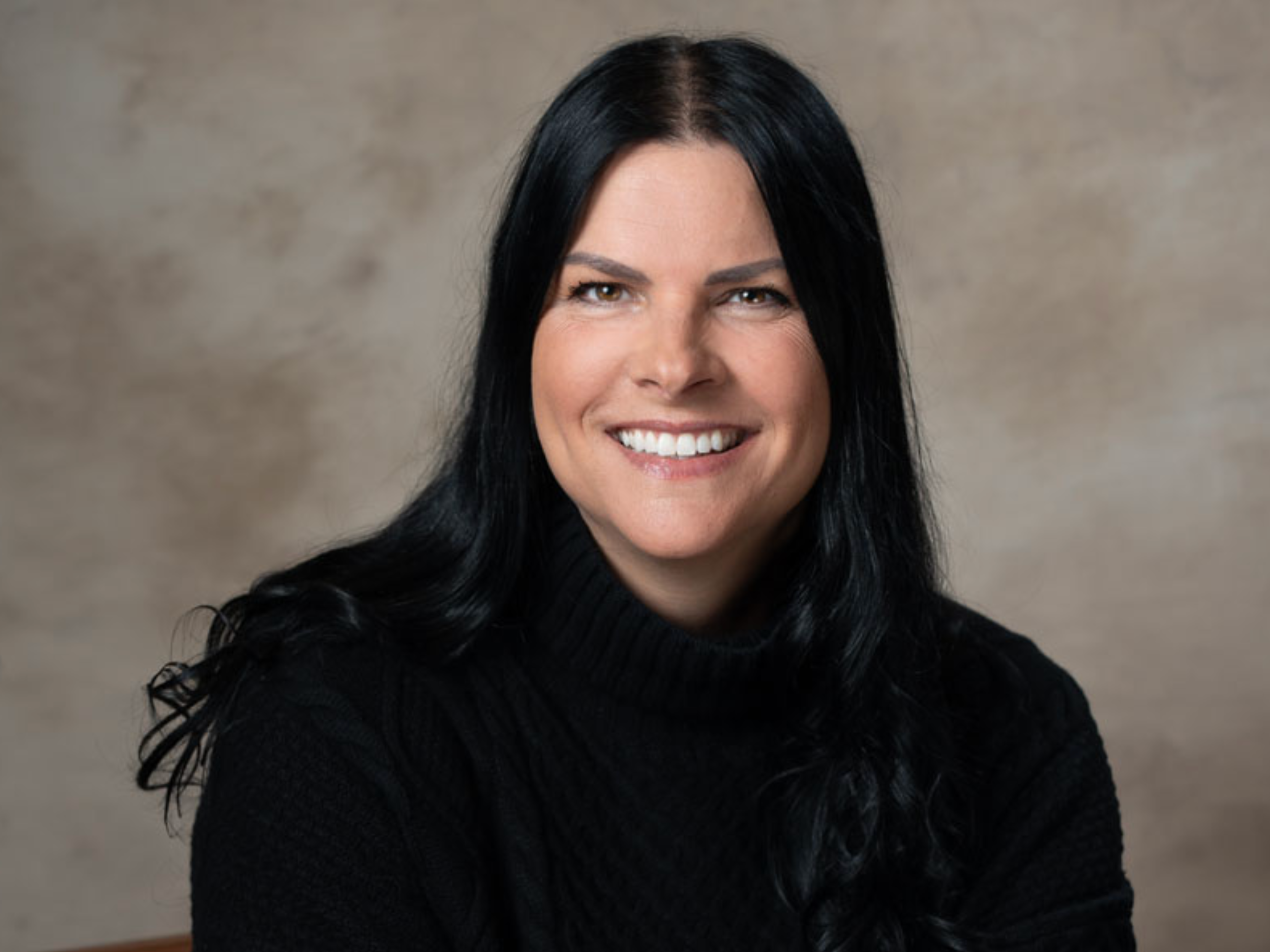 Smiling woman with long black hair wearing a black turtleneck sweater, portrait against a neutral background.