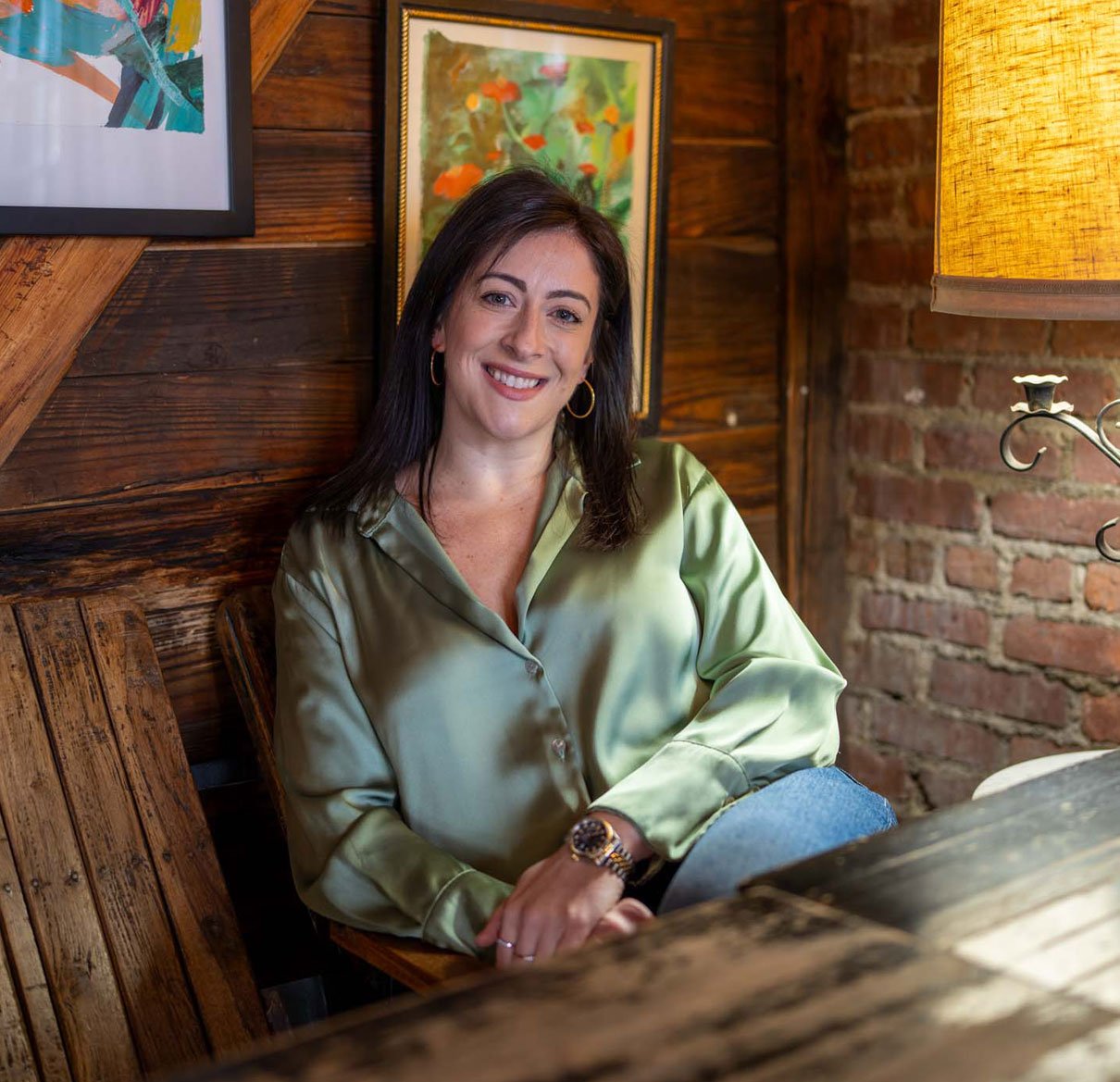 A woman with dark hair and hoop earrings smiling, sitting in a cozy room with wooden walls, a framed painting of flowers behind her, and a yellow lampshade next to her.