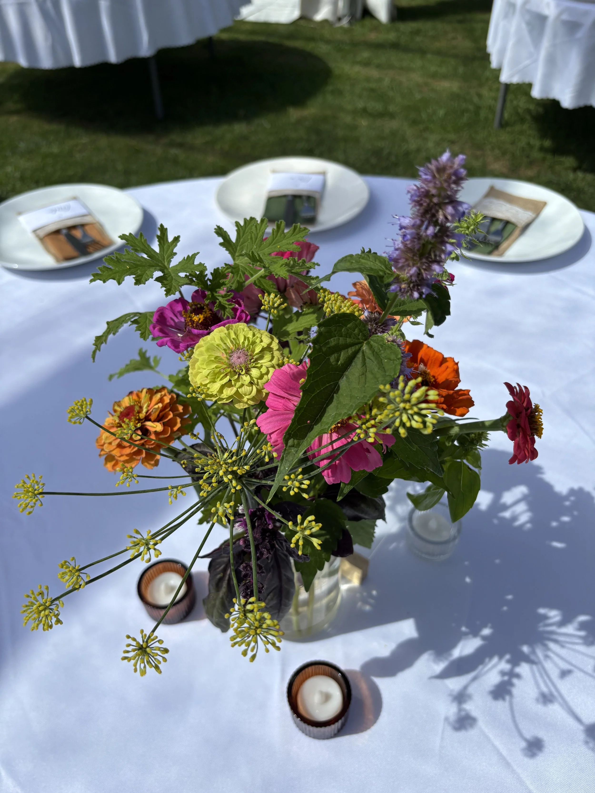 Colorful flower arrangement on a white tablecloth with three place settings and small tealight candles in brown holders