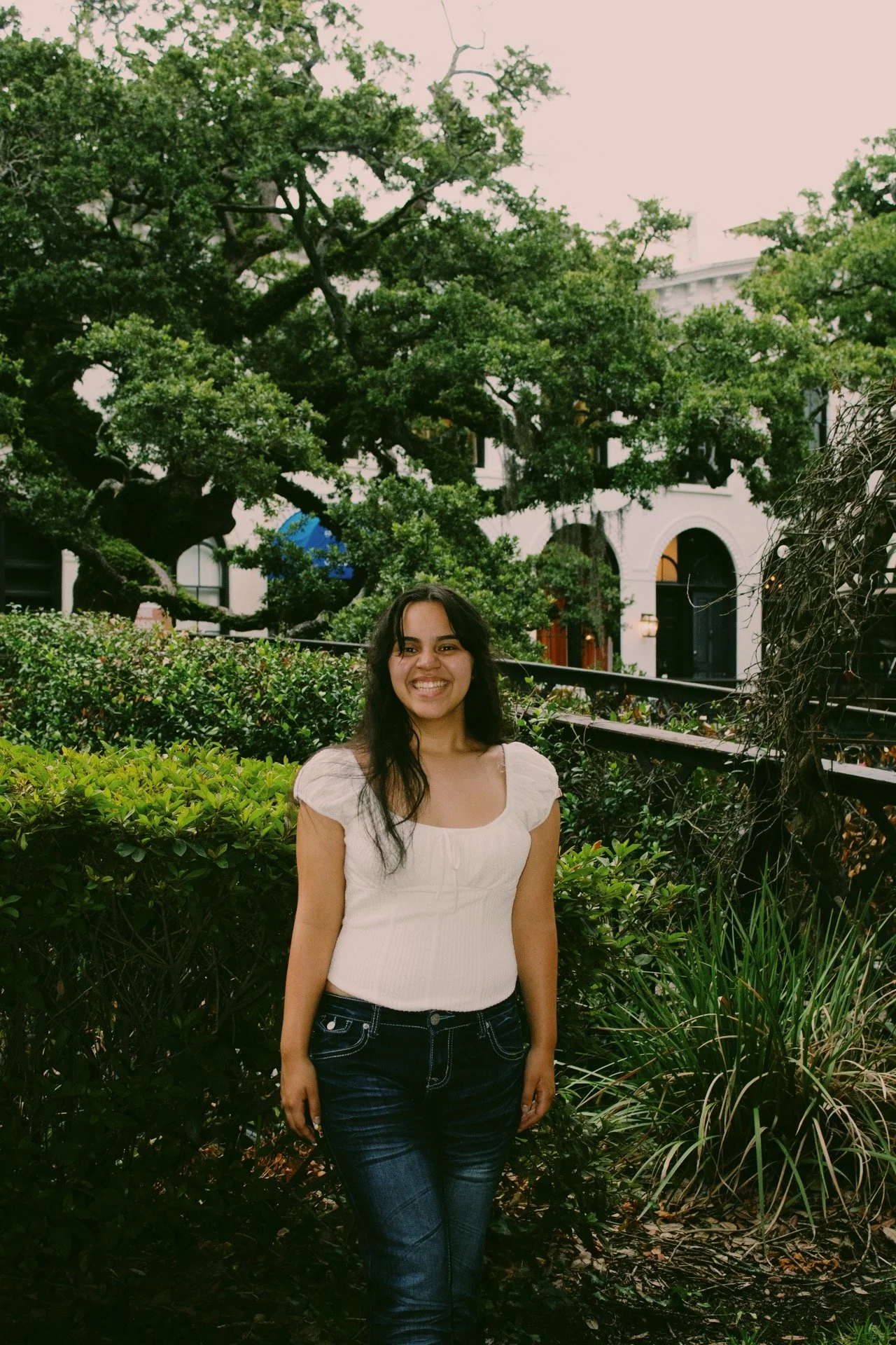 A young woman with long dark hair smiling in front of green bushes and trees, with a white building in the background.