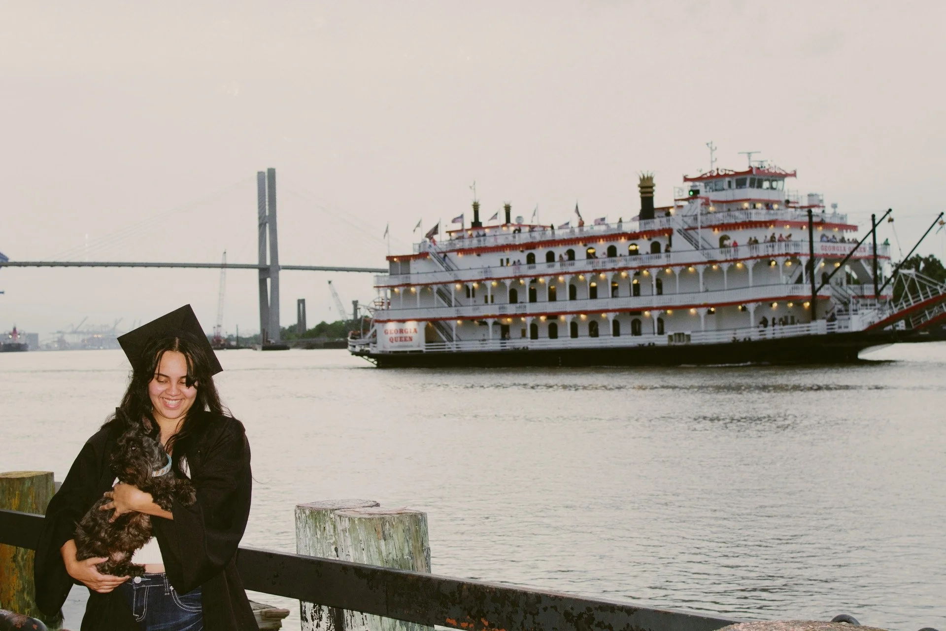 A woman in a graduation gown and cap smiling and holding a small dog, with a river, a large riverboat named Georgia Queen, and a bridge in the background.