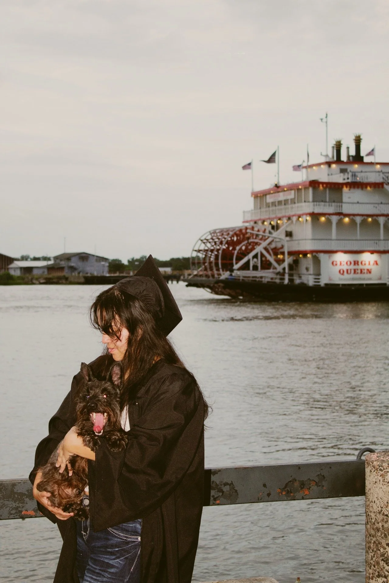 A woman in a graduation gown and mortarboard holding a small dog near a body of water, with a large paddle wheel riverboat named Georgia Queen in the background.