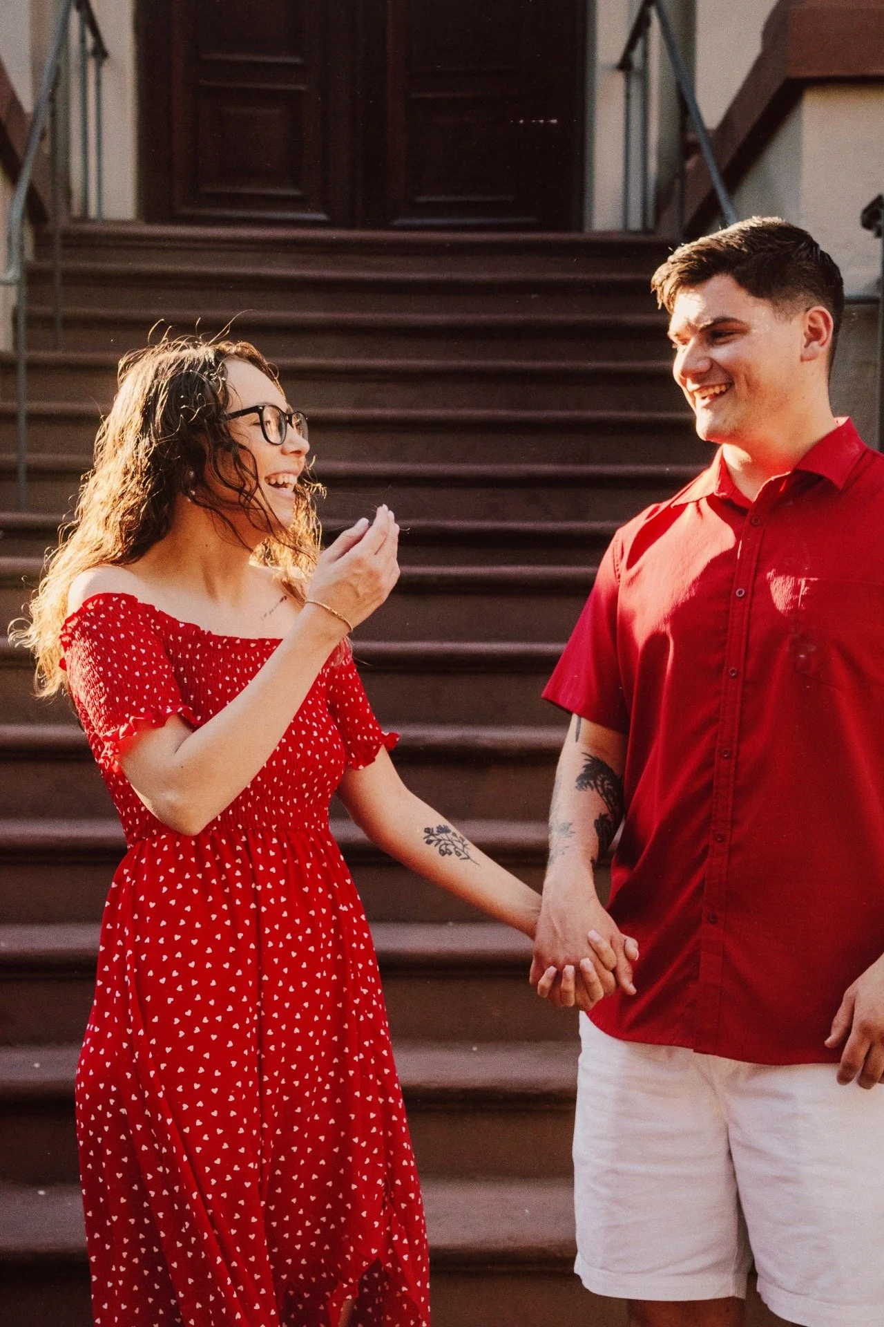 A woman in a red dress with white hearts and glasses holding hands with a man in a red shirt and white shorts on outdoor steps, smiling and laughing.