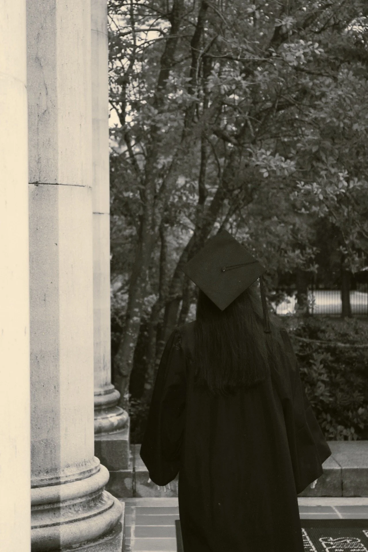 A person wearing a graduation gown and cap standing outdoors near stone columns and trees.