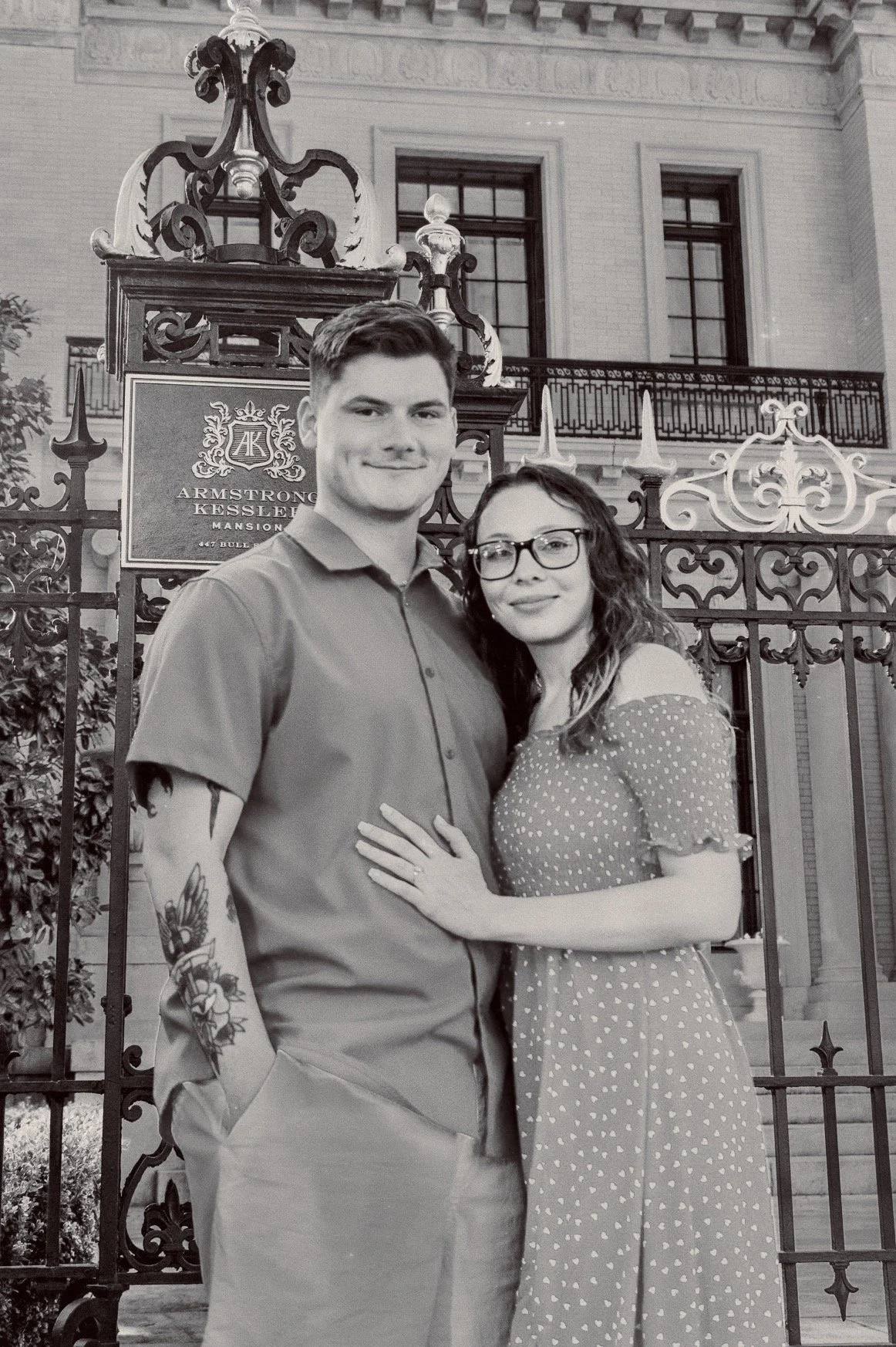 A young couple stands together in front of a historic building with ornate fencing and a sign that reads "Armstrong Kessler Mansion". The man is wearing a button-up shirt and has tattoos on his forearm. The woman is wearing glasses and a polka dot dr