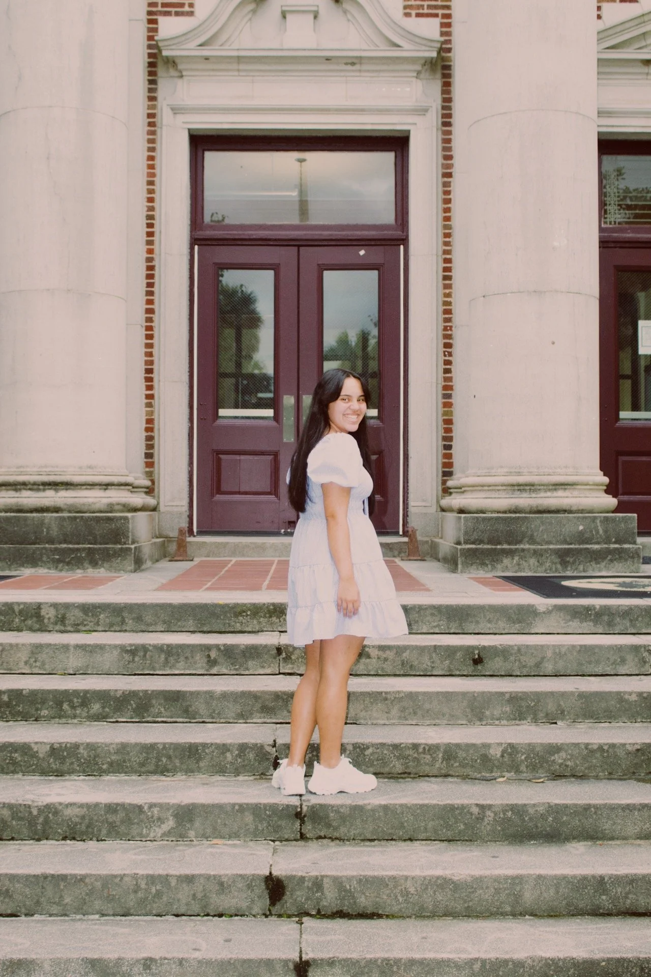 A young woman in a white dress and sneakers standing on stairs in front of a building with large columns and maroon double doors, smiling at the camera.