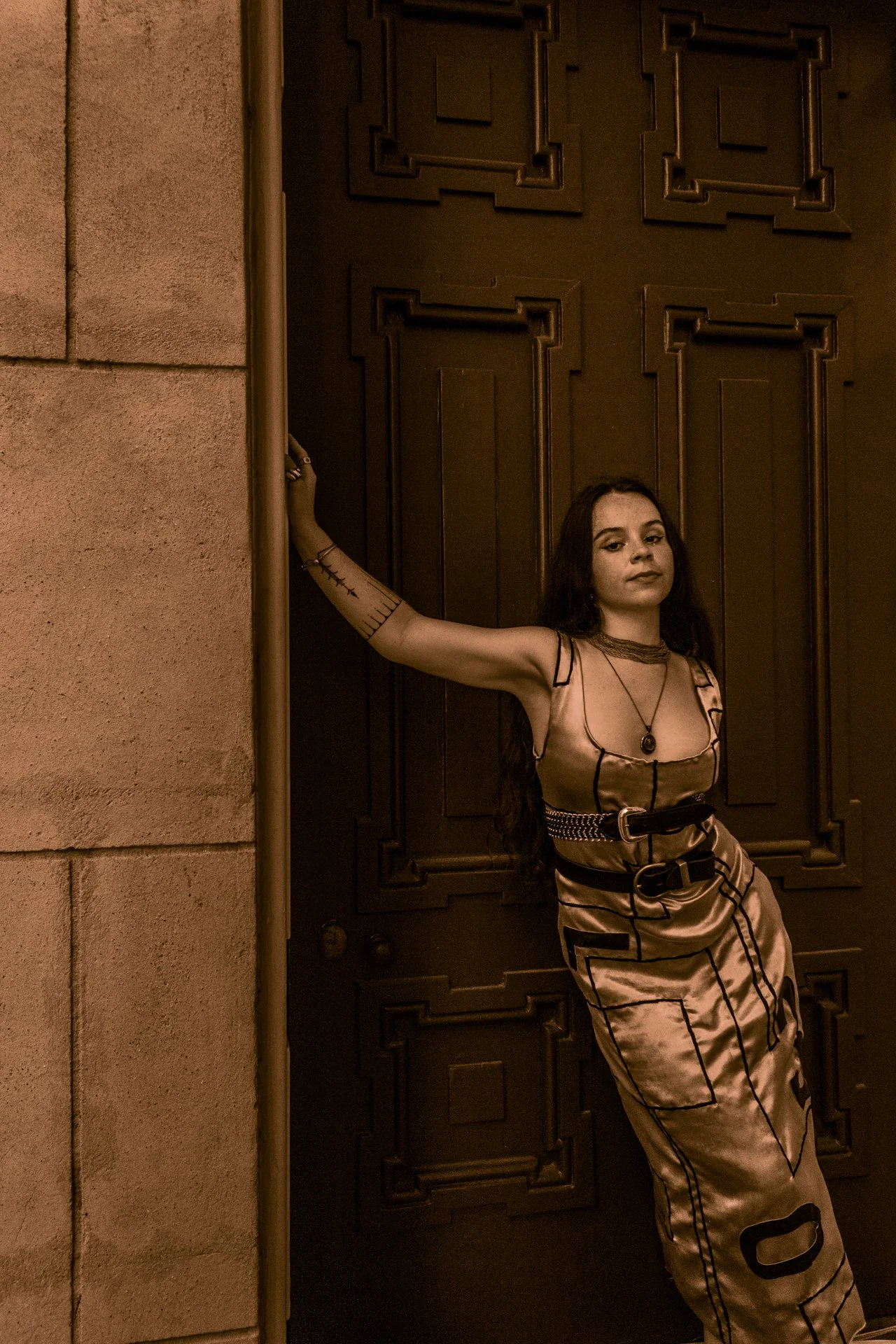 A woman with long dark hair and tattoos on her arm stands leaning against a wall and door, wearing a satin dress with black lines and a belt, accessorized with necklaces, in a dimly lit interior setting.