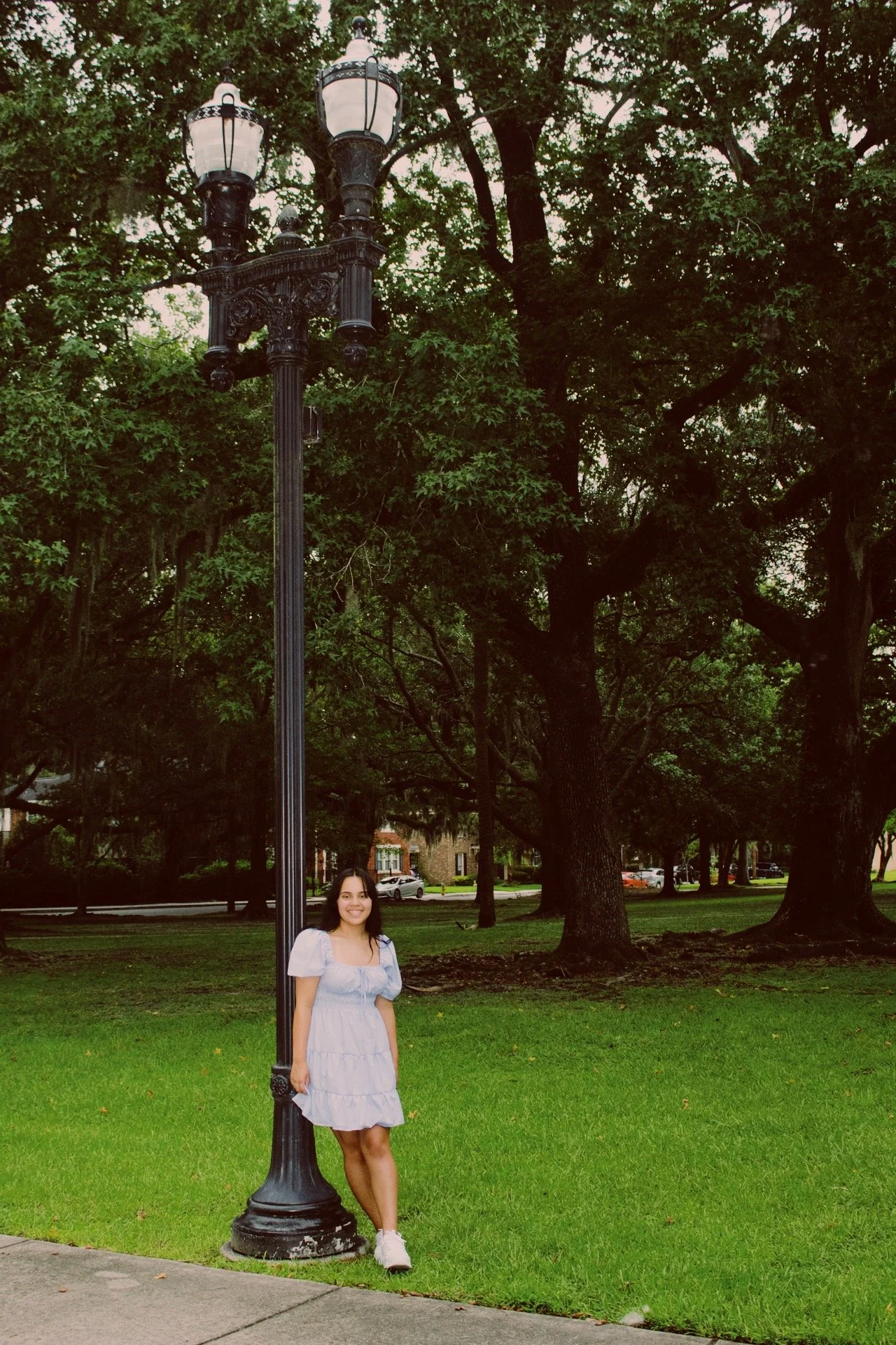 A young woman in a white dress and sneakers standing next to a black street lamp in a park with green grass and large trees in the background.