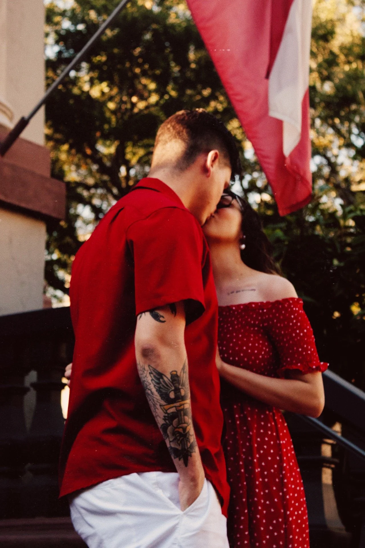 A young couple sharing a kiss outdoors, with the man wearing a red shirt and white pants, and the woman in a red polka dot dress. There is an American flag visible in the background.