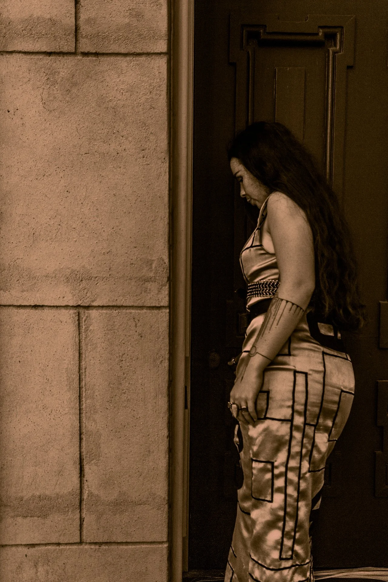 A woman with long dark hair wearing a satin top and matching pants, standing with her head bowed against a door behind her, in sepia tone.