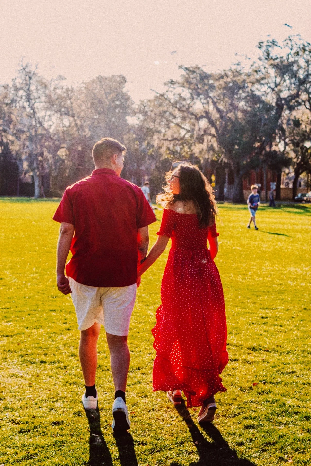 A young couple walking hand in hand on a sunny grassy field, with trees and other people in the distance.