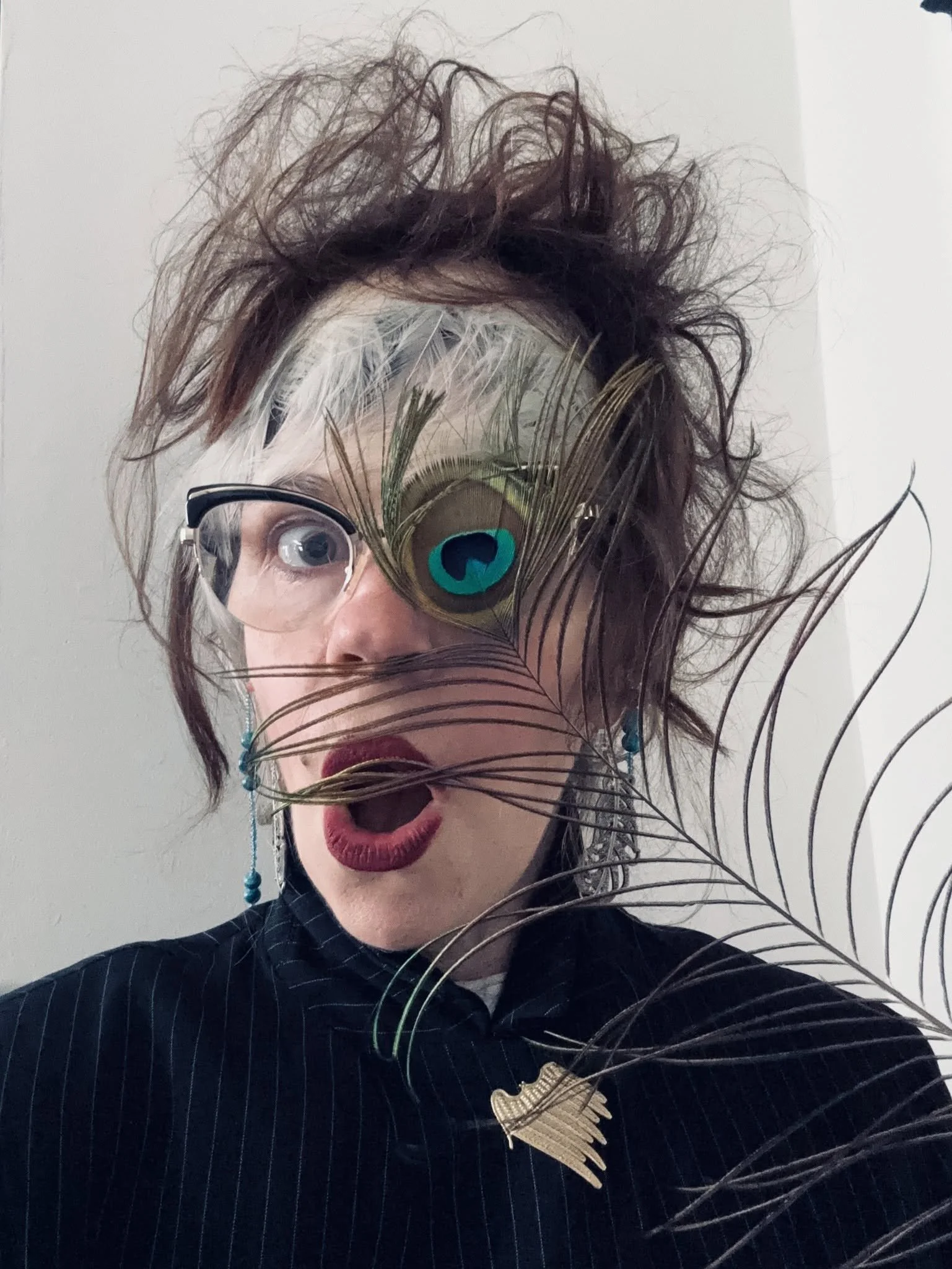 A person with messy, curly hair, wearing glasses, red lipstick, and a dark pinstripe suit, holding a peacock feather in front of their face.