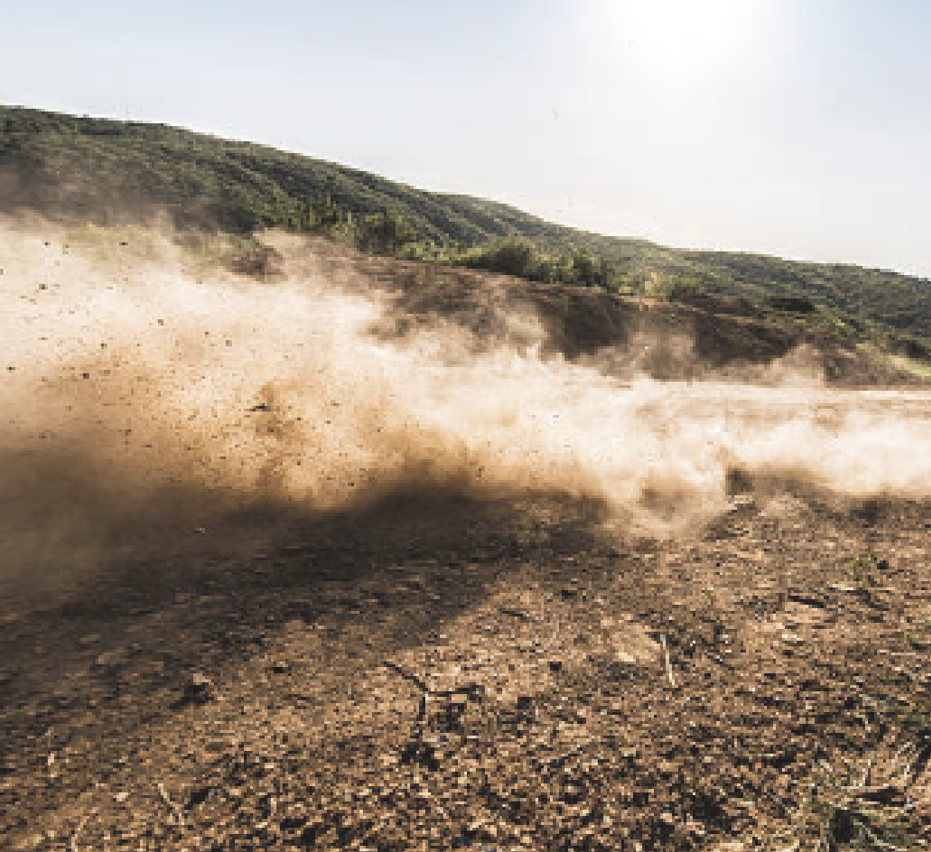 Dust storm blowing over a rocky hillside with green trees in the background.