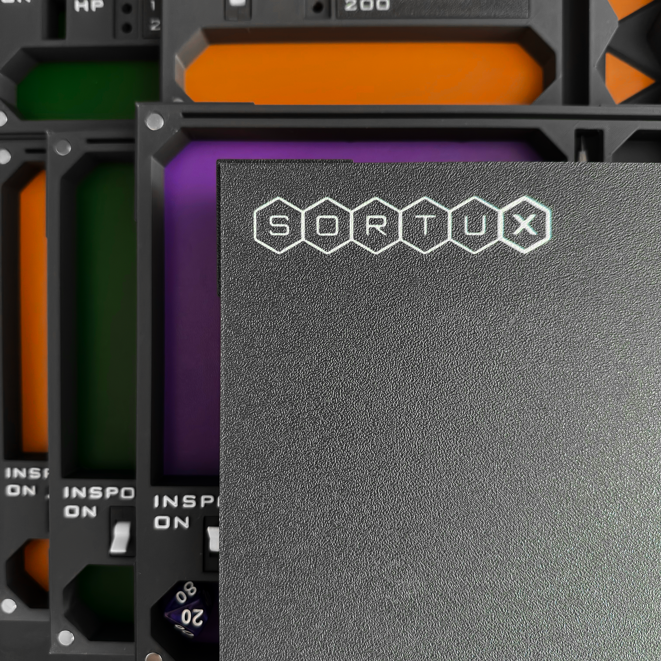 Close-up of a black covered gaming or tech device labeled 'SORTUX' resting on colorful dice trays.