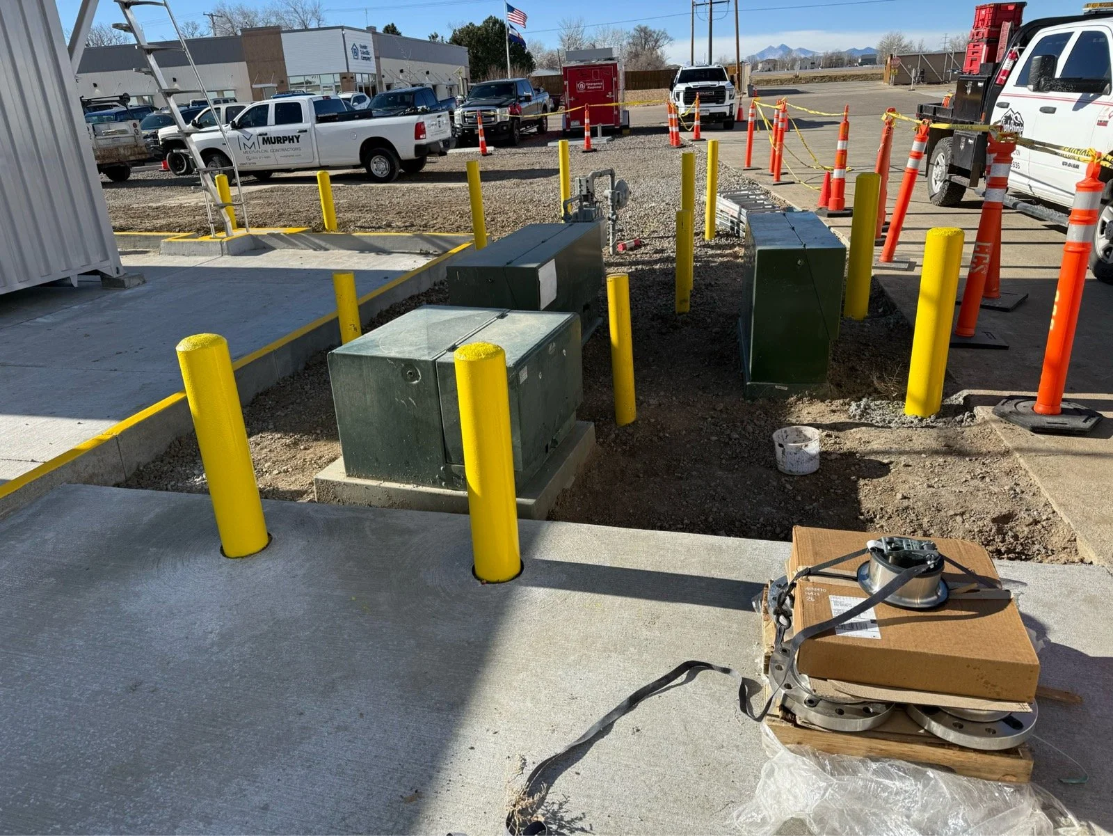 Construction site with electrical boxes, yellow bollards, orange traffic cones, and parked pickup trucks in the background under a clear sky.