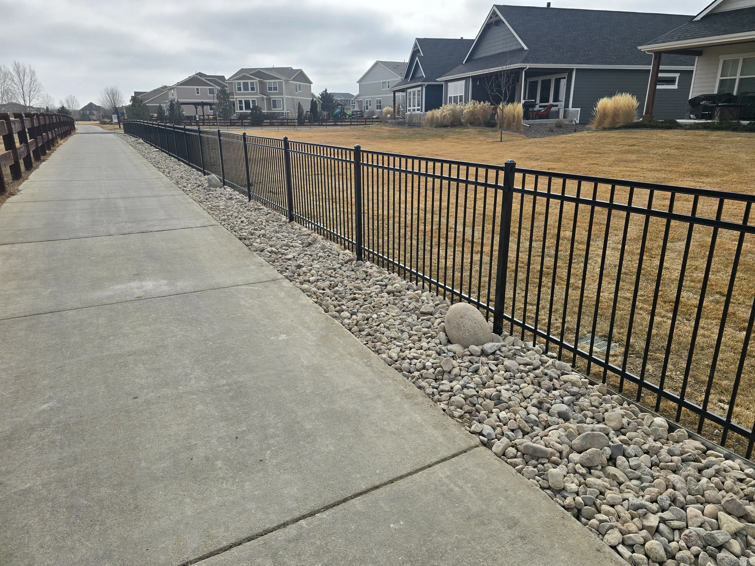 Sidewalk with black metal fence running alongside a row of suburban houses with well-kept lawns.